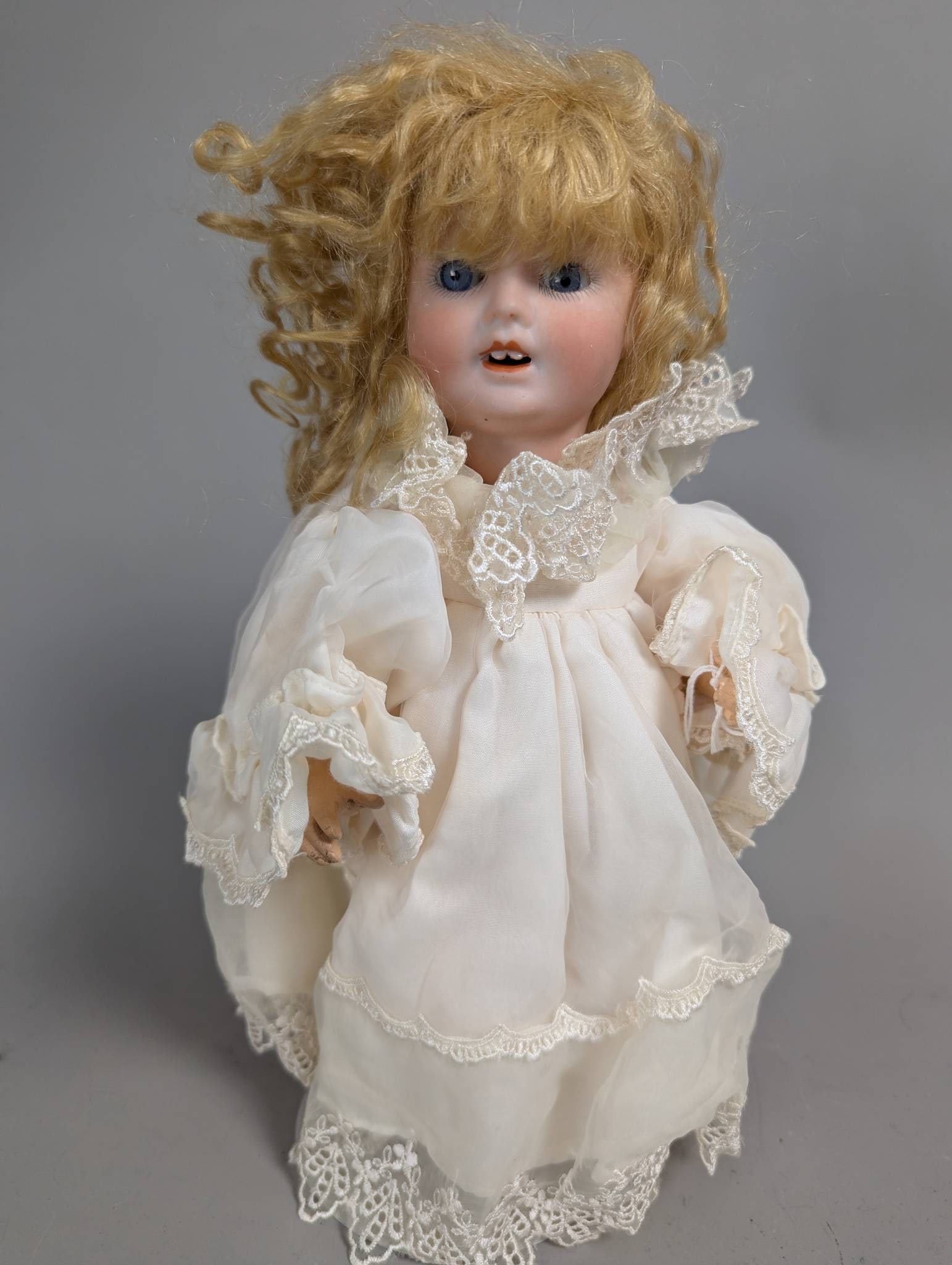 Bisque Head Doll with Blonde Hair and Lace Dress: Bisque head doll measuring 13 inches high, featuring blonde hair and blue eyes with an open mouth. The doll is dressed in a delicate lace-trimmed white dress. The bisque head shows details in the faci