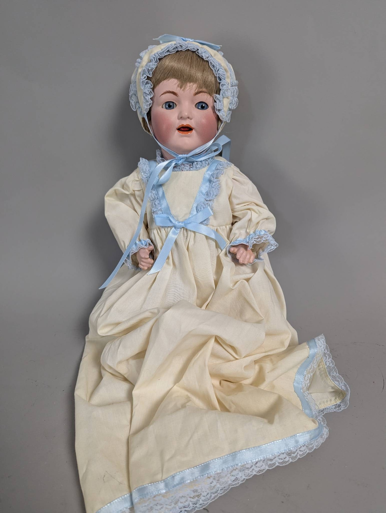 Simon and Halbig 126 Bisque Head Doll Dressed in Bonnet and Gown: Simon and Halbig 126 bisque head doll, standing 17 inches high, featuring hand-painted facial details with blue glass eyes and an open mouth showing teeth. The doll wears a cream gown accented with bl