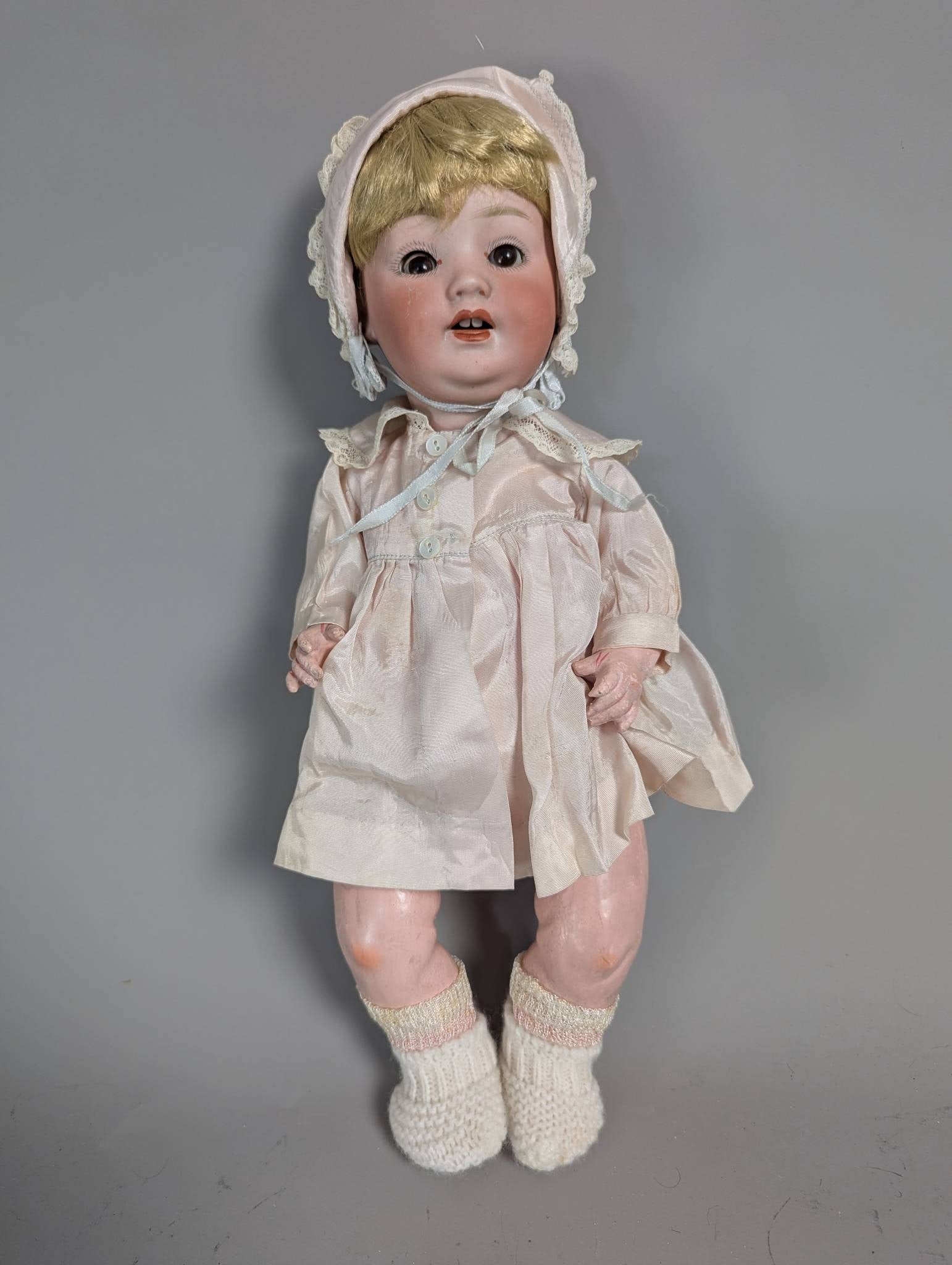15 Inch Doll by Puppen- und Spielwarenfabrik MOM: Vintage 15-inch doll by Puppen- und Spielwarenfabrik MOM featuring a bisque head with painted facial details, synthetic blonde hair, and fixed eyes. The doll is dressed in a delicate pink dress with l