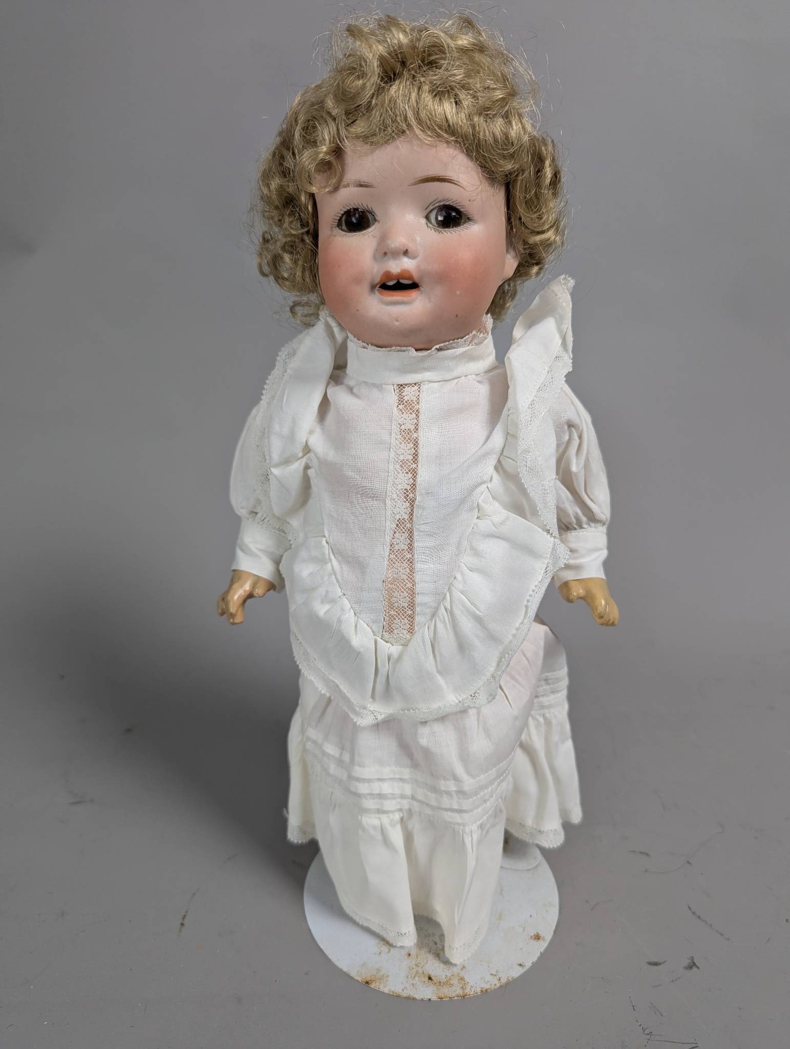 Porcelain Doll by Herm and Steiner with White Dress and Blonde Curls: Porcelain doll attributed to Herm and Steiner, measuring 12 inches high. Features a bisque head with hand-painted facial details, including glass eyes and an open mouth with teeth. The doll is adorned