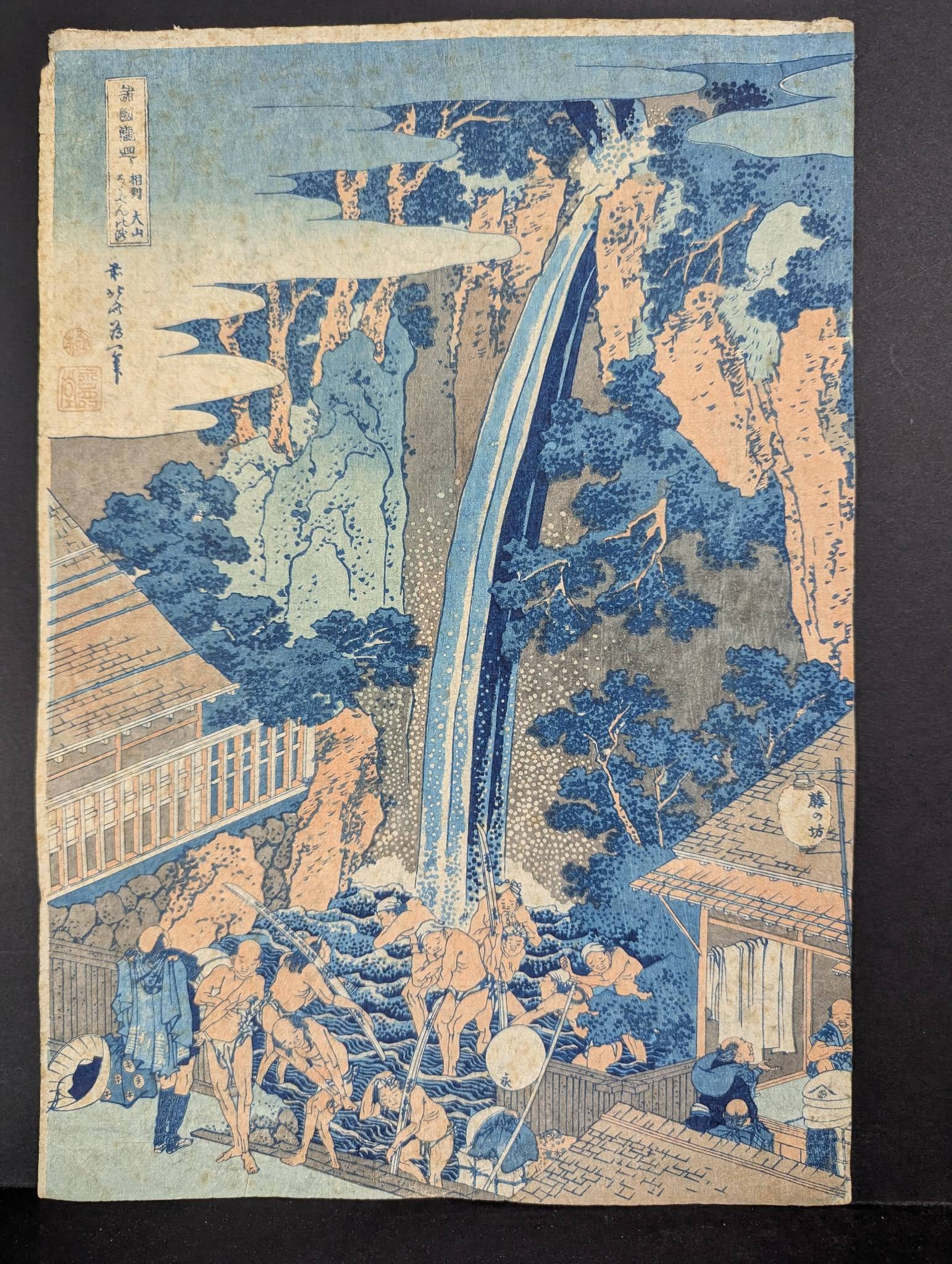 Japanese Woodblock Print Reproduction by Hokusai Depicting a Waterfall Scene: Reproduction Japanese woodblock print by Hokusai featuring a vibrant waterfall scene. The artwork measures 10 inches high by 14 3/4 inches wide. Visible signs of age include foxing, tape residue on th