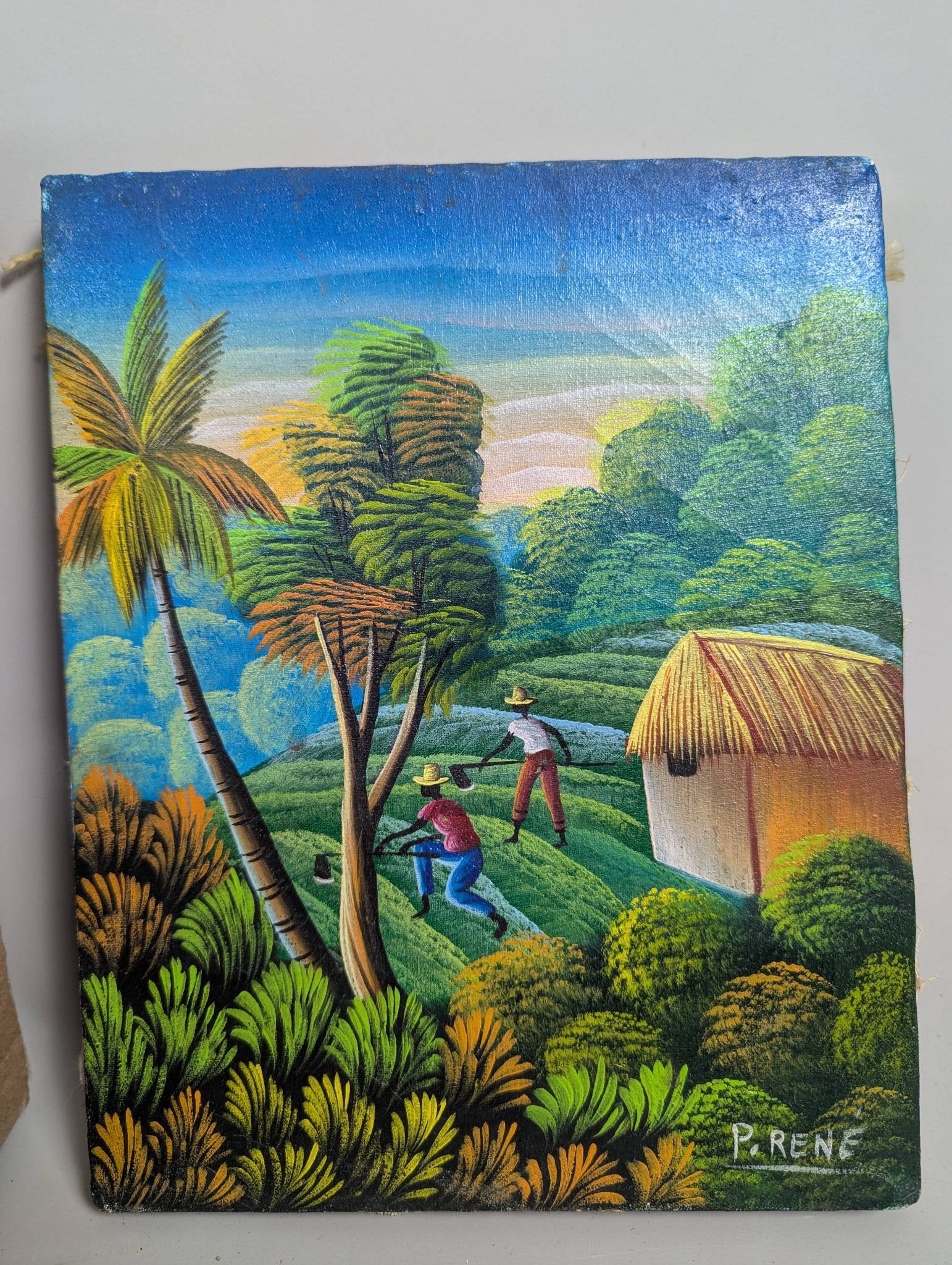 Haitian Folk Art Oil Painting Depicting Tropical Village by P Rene: A Haitian folk art oil painting signed by P Rene. The artwork depicts a vibrant tropical village scene showcasing lush greenery, a thatched-roof hut, palm trees, and two figures working in a field