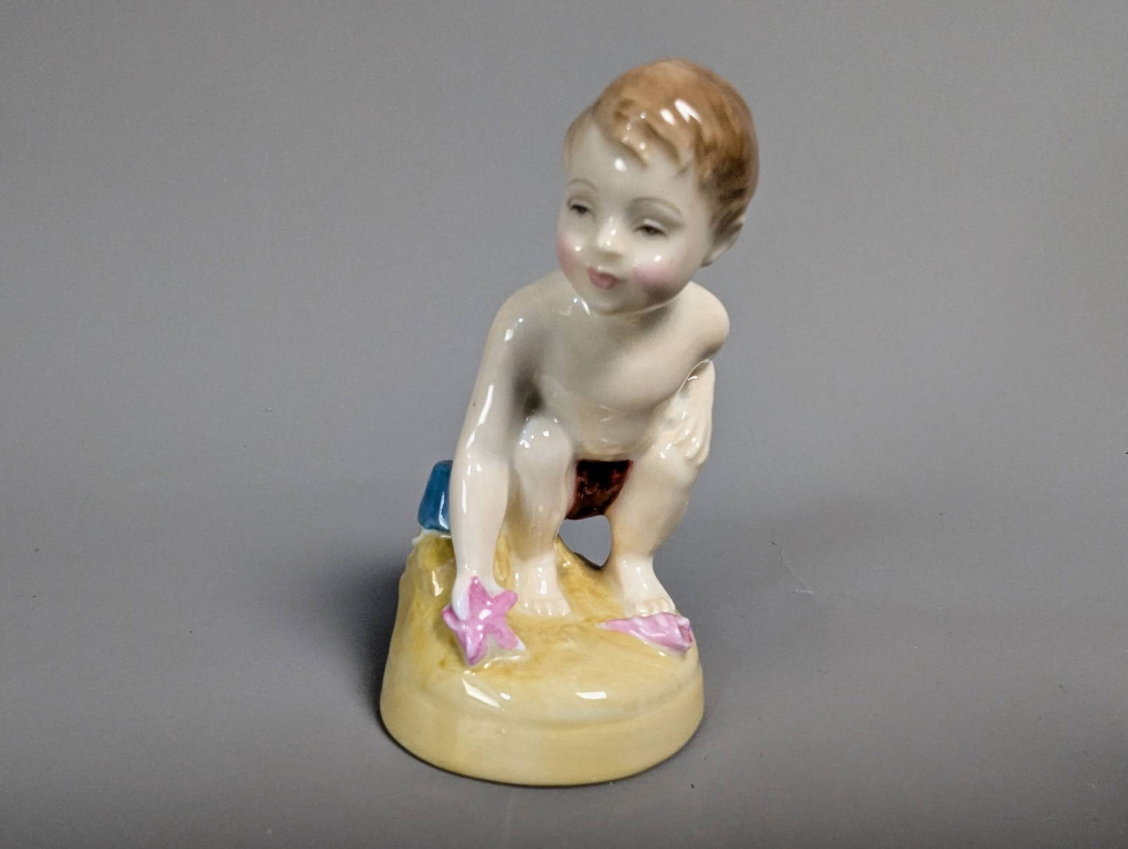 Royal Doulton HN2261 Sea Shore Ceramic Figurine: Royal Doulton HN2261 Sea Shore figurine, handcrafted in fine bone china, dated 1960. Depiction of a young boy crouching on the sand with a pink starfish and beach toys around him. The piece is meticul
