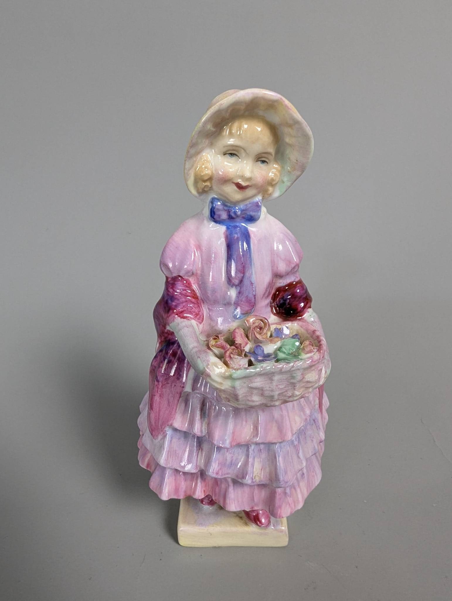 Royal Doulton Greta Porcelain Figurine in Purple Dress: Royal Doulton porcelain figurine titled Greta. The figurine features a woman in a vintage-style purple dress and bonnet, holding a woven basket of colorful flowers. Marked with the Royal Doulton backs