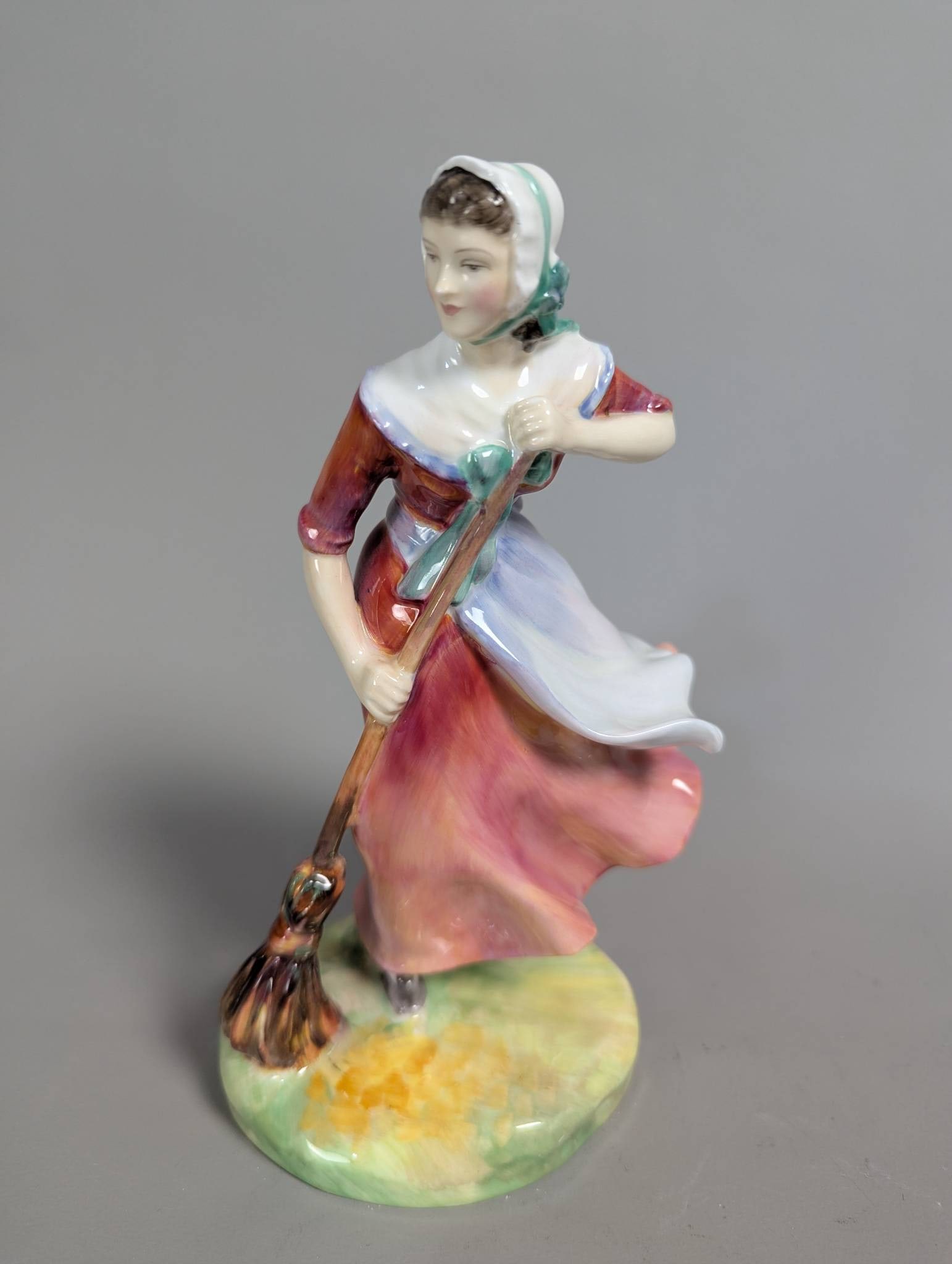 Royal Doulton hn 2087 Autumn Porcelain Figurine: Royal Doulton hn 2087 Autumn porcelain figurine. This collectible figurine stands 7 1/2 inches high and features a woman in a colorful dress with a sweeping pose and a broom, set on a green and yellow