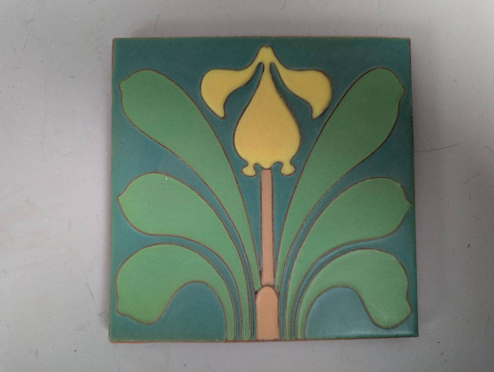 American Van Briggle Arts and Crafts Style Pottery Tile with Iris Motif: American Van Briggle Arts and Crafts style pottery tile featuring an iris or tulip motif. The tile has been hand-glazed in a matte finish, showcasing harmonious yellow, green, and blue tones. The desi
