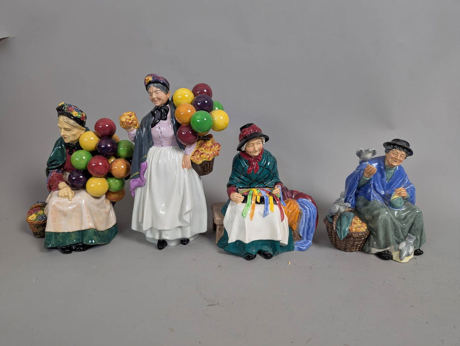 Royal Doulton Figurines Including Old Balloon Seller and Tuppence a Bag: Four Royal Doulton porcelain figurines, including Old Balloon Seller HN1315, Biddy Pennyfarthing HN1843, Silks and Ribbons HN2017, and Tuppence a Bag HN2320. Each hand-painted figurine features