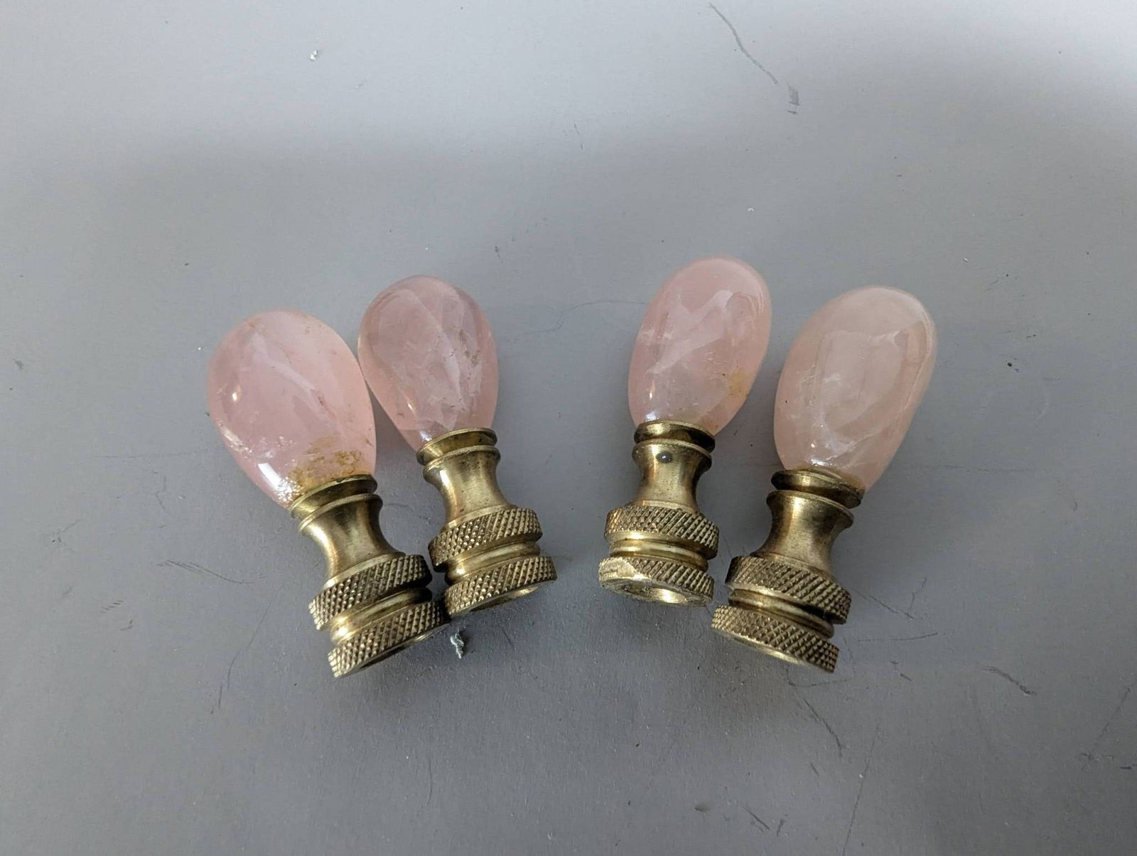 Four Rose Quartz Lamp Finials with Brass Bases: Four rose quartz lamp finials featuring brass bases with a textured grip design. Each finial measures 2 inches high by 1/2 inch wide by 1/2 inch deep. The rose quartz elements offer a subtle pink hue
