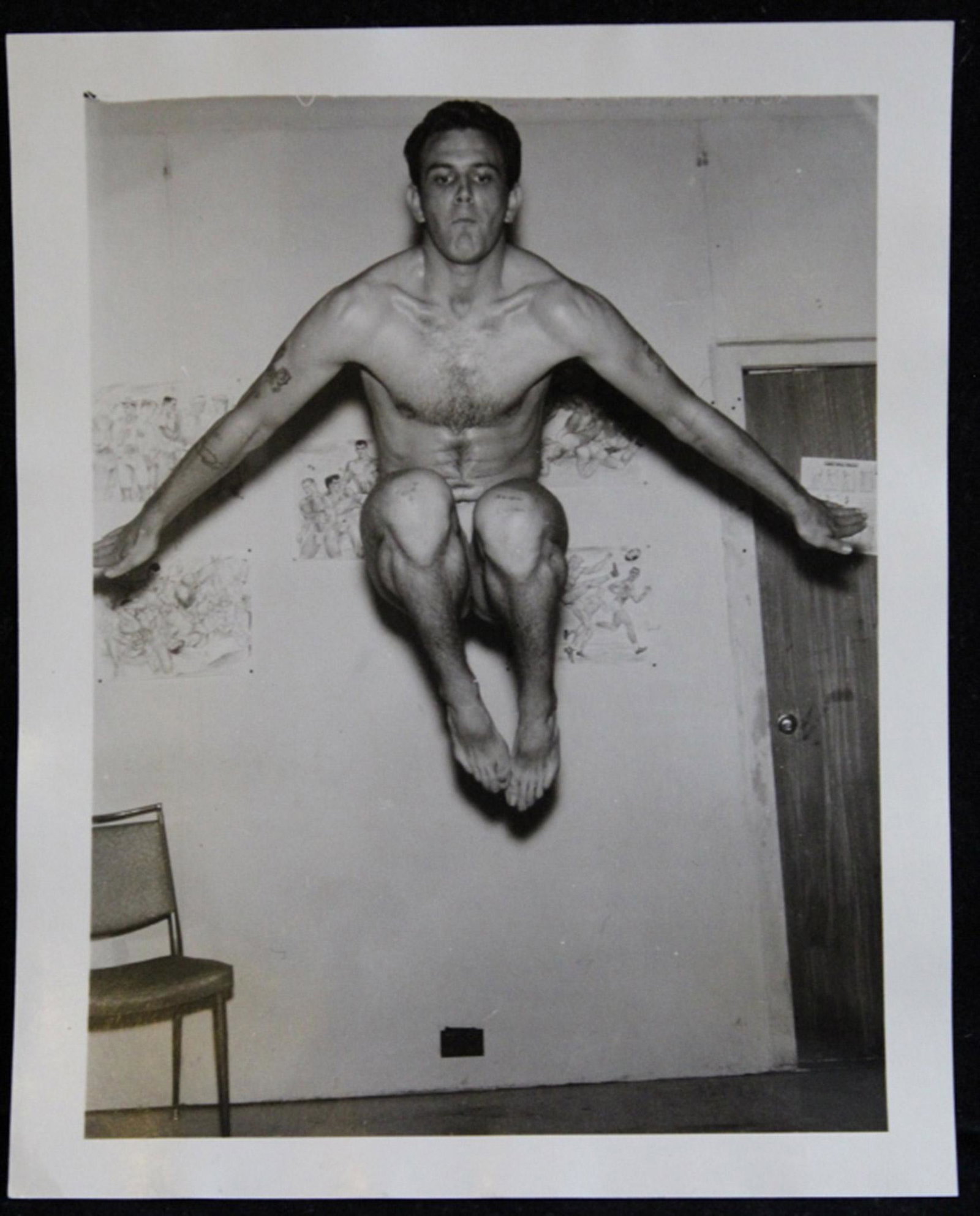 Bob Mizer AMG Male Semi Nude Photograph: Tattooed model leaping in air in front of homoeroctic drawings pinned to the wall. Pencil numbered 39 to reverse. Good sharp image printed on semi-gloss paper. Circa 1950's. 4" x 5".