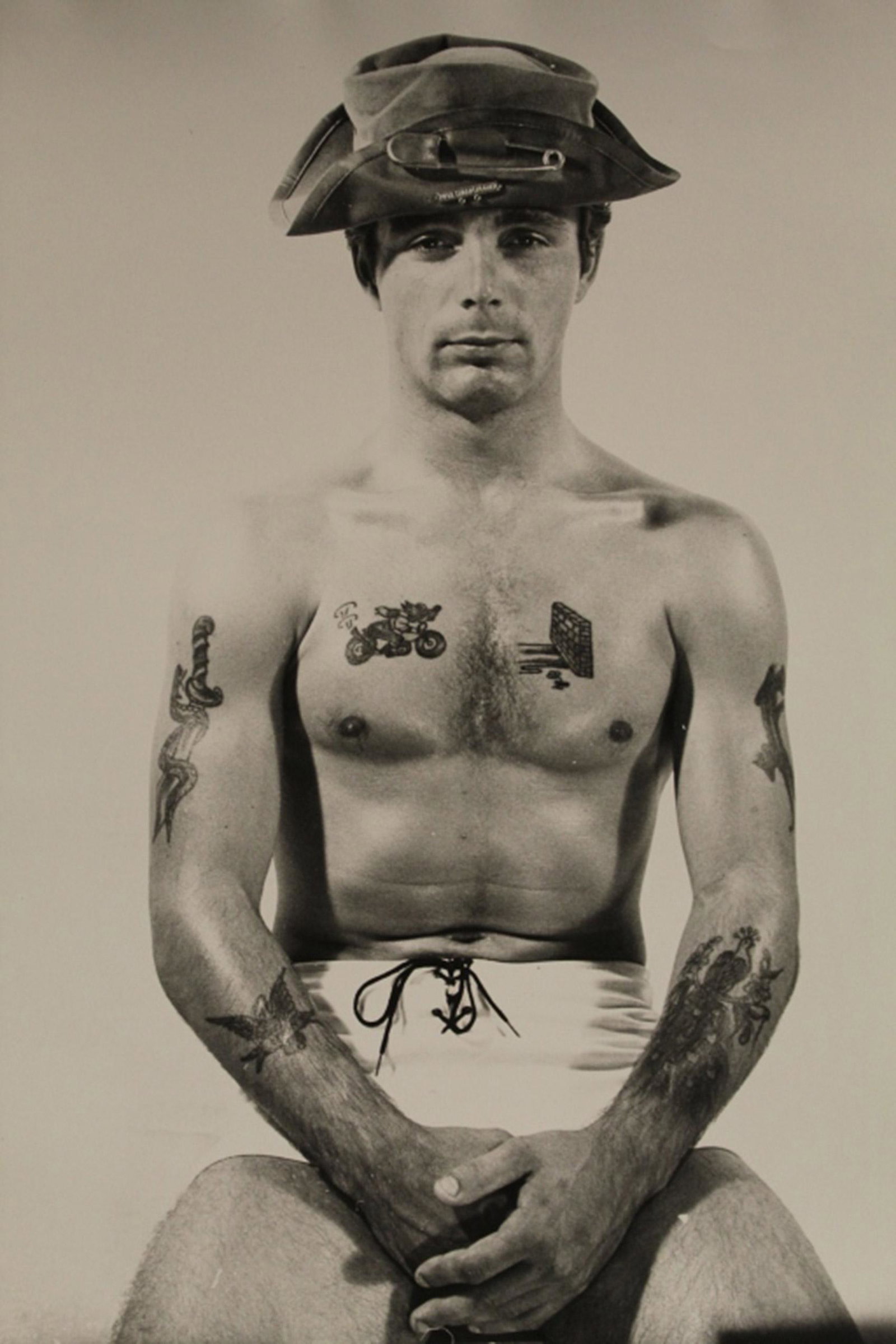 AMG Bob Mizer Semi Nude Male Photograph: Shirtless man with chest and arm tattoos wearing small lace up shorts and hat. Excellent clear sharp image on single weight satin paper. Circa 1960's/ 70's. 8" x 10".