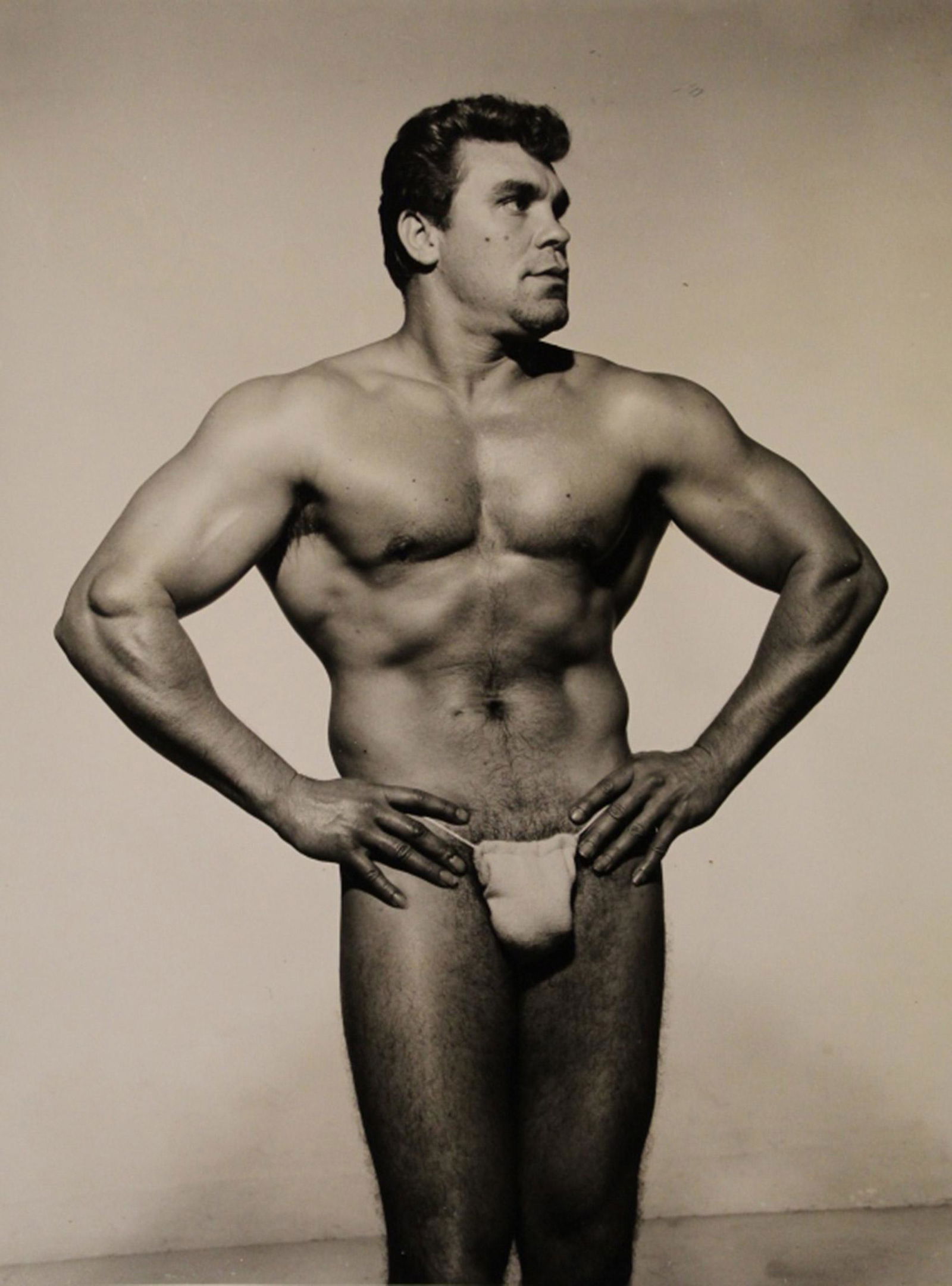 AMG Bob Mizer Male Semi Nude Photograph: Male bodybuilder in small posing strap with hands on hips. Pencil written 9 to reverse; glue / paper residue spots (does not affect image). Excellent clear sharp image on gloss paper. Circa 1950's. 4"