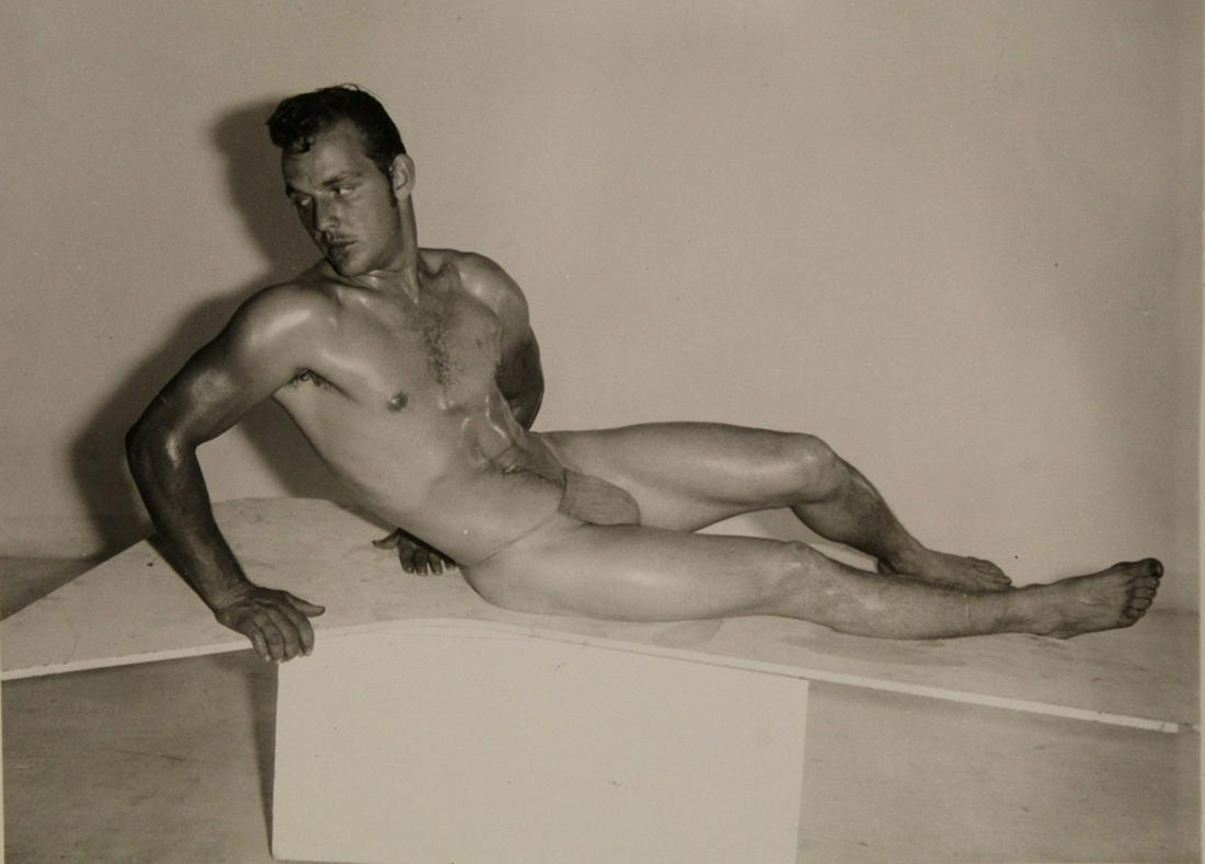 AMG Bob Mizer Male Semi Nude Photograph: Model posed on geometric platform bench wearing posing strap. Pencil written 22 to reverse. Small light corner fold upper left. Excellent clear sharp image on gloss paper. Circa 1950's. 4" x 5".