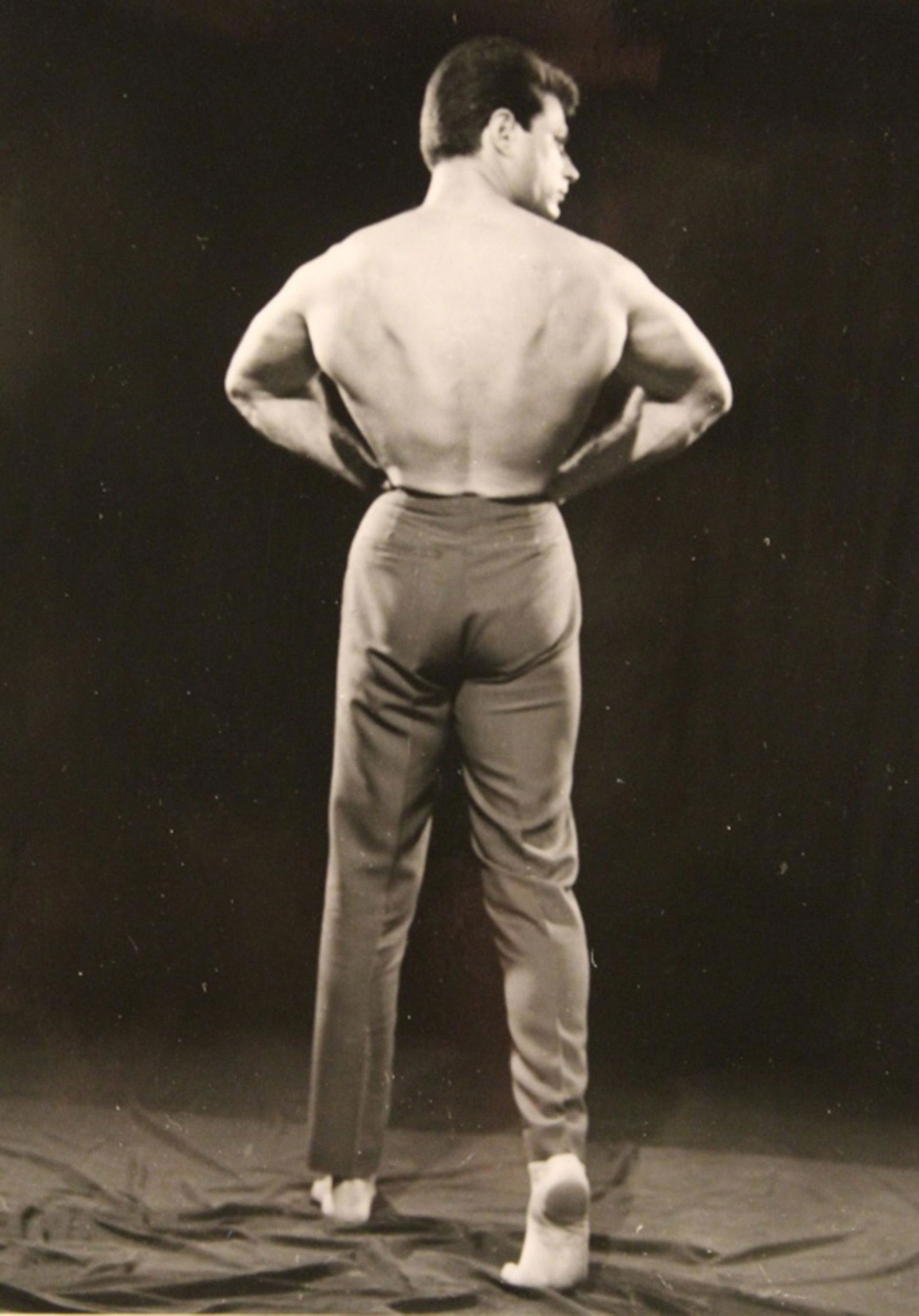 Vintage Bob Mizer AMG Semi Nude Male Photograph: Bodybuilder performing a rear lat spread back pose while wearing suit pants. Pencil written "12" to reverse. Excellent clear sharp image on gloss paper. Circa 1950's. 4" x 5".