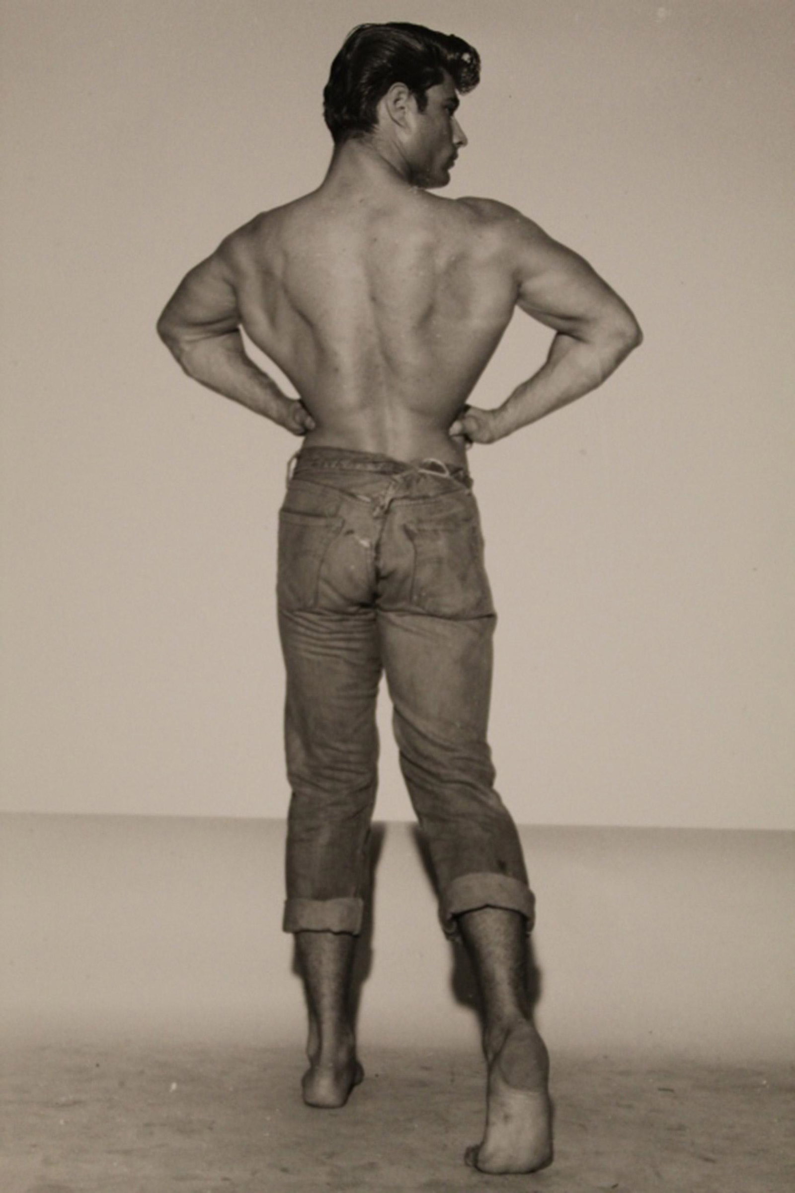 Bob Mizer AMG Male Semi Nude Photographs: Two photographs of male bodybuilders performing rear lat spread poses (one wearing jeans and one wearing dress pants). Excellent condition printed on gloss paper (YR11-AM and 12 pencil written notatio