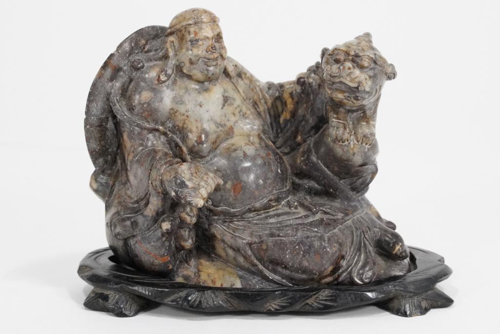 Chinese Carved Soapstone Buddha Figure: Reclining Buddha figure with hand on foo dog/ lion. Old repaired break (with small loss) to large hat behind head. No apparent artist mark. Includes custom carved hardwood stand. 4.5" H x 5.5" L x 3.5