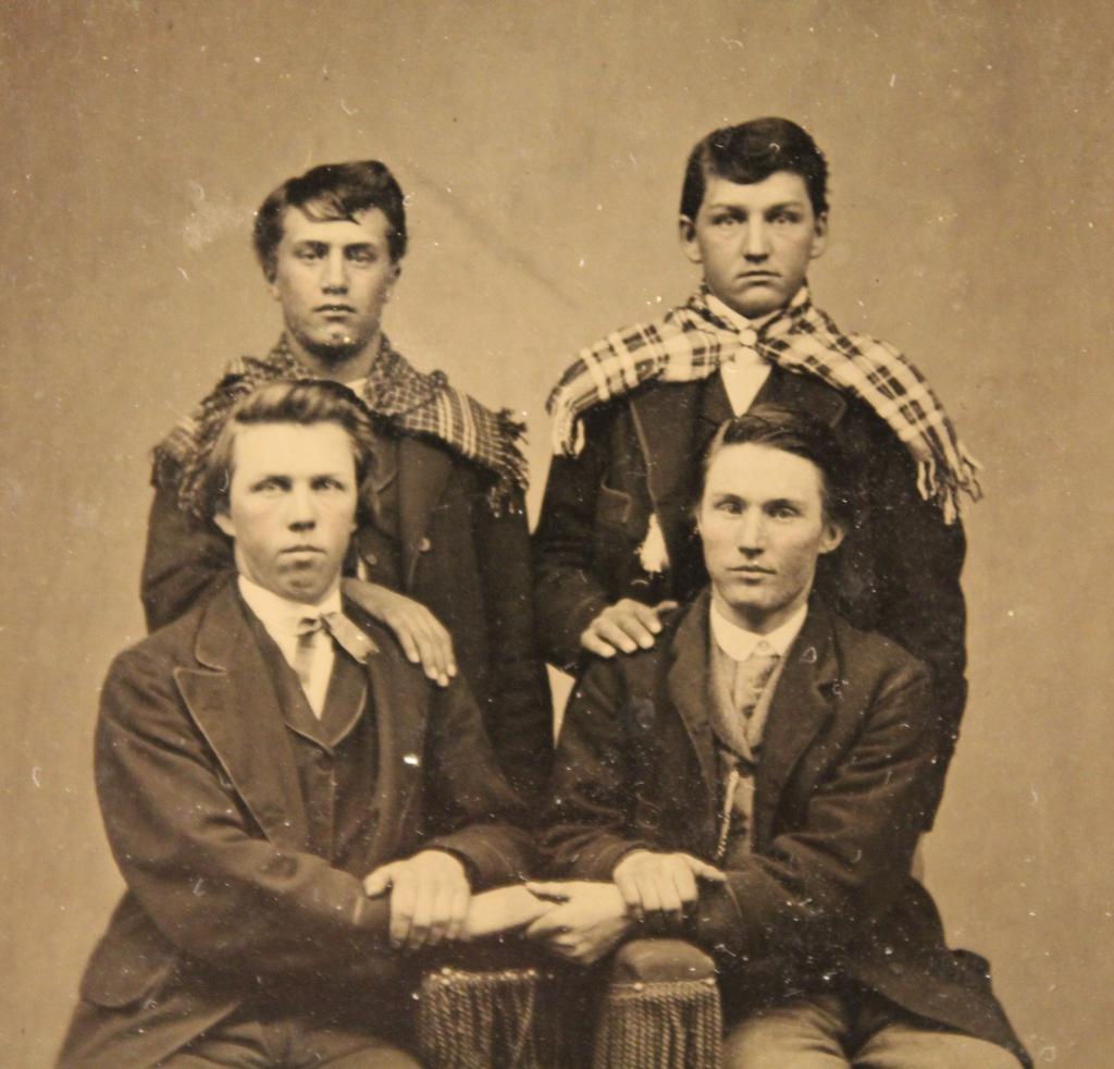 Antique Tintype Photograph of Affectionate Men: Studio tintype of four young men; two men posed standing (wearing plaid scarves) with their hands resting on the shoulders of two men posed sitting intimately holding hands. Clear and sharp image; nor