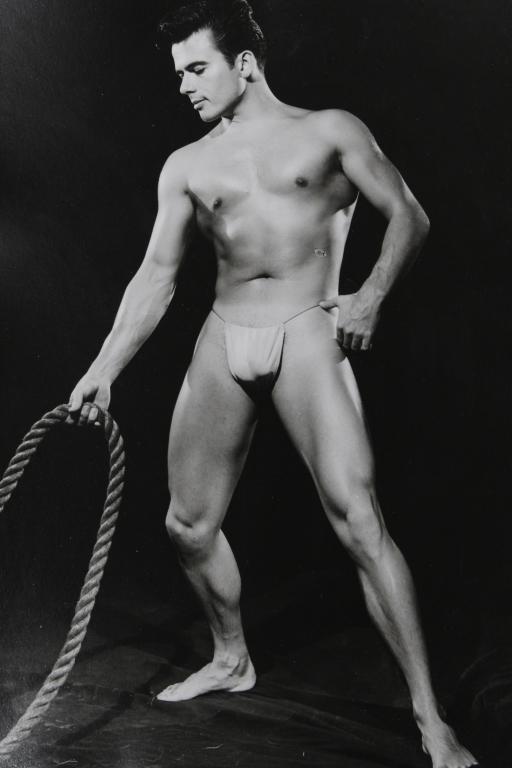 Bob Mizer AMG Male Nude Photograph: Male semi-nude model in studio posed with large rope. Hand written "XGII-L" to the reverse. Clear sharp silver gelatin print on satin heavy fiber paper. Circa 1950's. Excellent condition with small ne