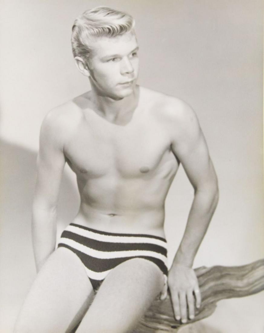 Bruce of LA / Bob Mizer Male Nude Photograph: Blonde semi nude model posed in studio wearing striped bathing suit sitting on a piece of gnarled wood. No identifying stamps. Slight sepia tone silver gelatin print on heavy fiber paper. Slight soft