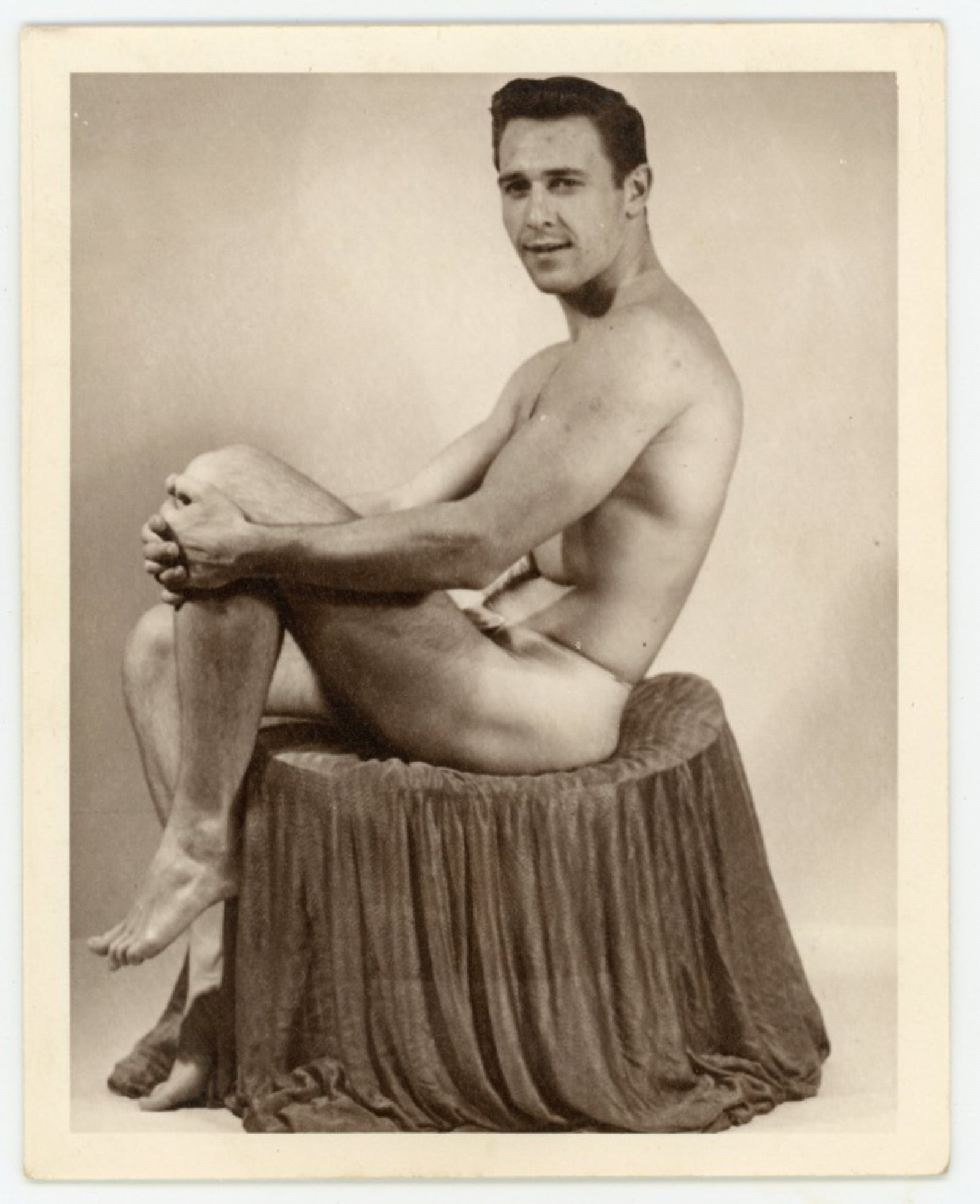 Kris Studio Male Physique Beefcake Photograph: Vintage original 4" x 5" Chuck Renslow / Kris Studio photograph of nude male model posed on fabric draped stool. Stamped "KRIS STUDIO P.O. BOX 1251 CHICAGO 90, ILL." in red to the reverse. Slight sepi