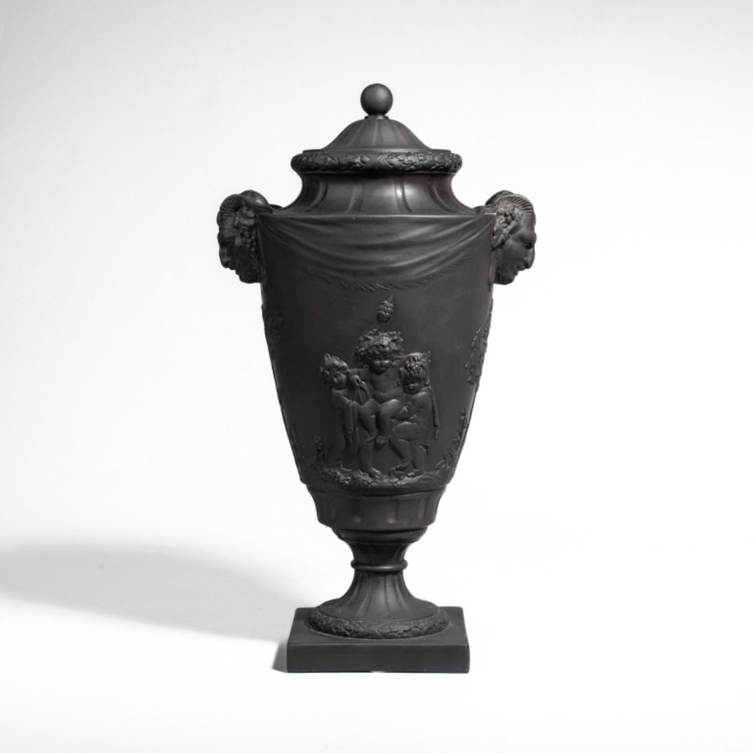 Wedgwood Basalt Urn Satyr Handle Urn / Cherubs: 19th century Wedgwood black basalt urn with lid. Satyr head handles with molded drapery between. Raised cherub and satyr figures to each side. Impressed "WEDGWOOD" to bottom. Two piece urn connected w