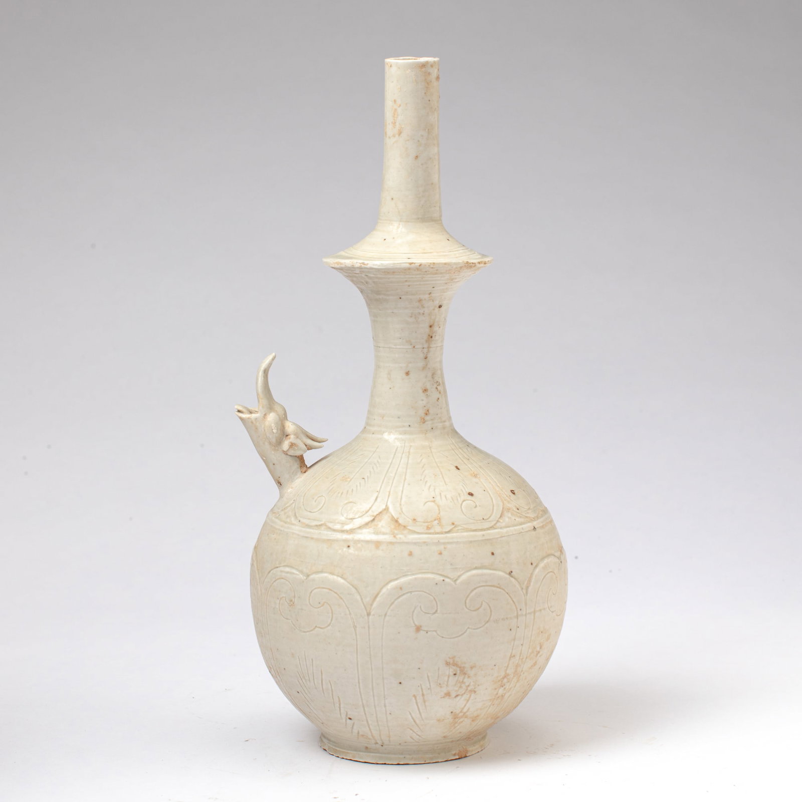 Chinese Ding-Type White Glazed Kundika: Chinese Ding-type white-glazed water sprinkler / kundika, having dragon-form spout above and within stylized petals in relief. Small loss to dragon; some fritting to rim and a few small firing flaws. 