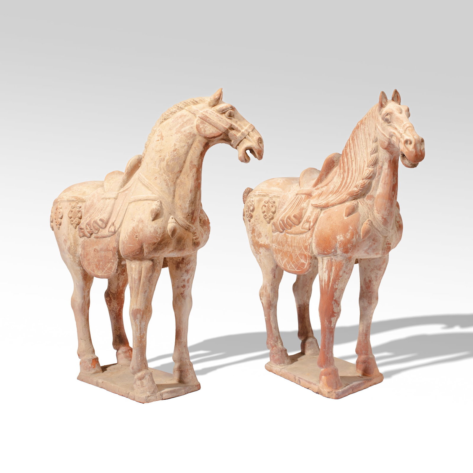 Pair Large Chinese Pottery Model Caparisoned Horse: Pair of Large Chinese models of caparisoned horses in red pottery with traces of slip-based pigment and modeled applique elements to body. One horse with loss to tail and base; the other has tail re-g