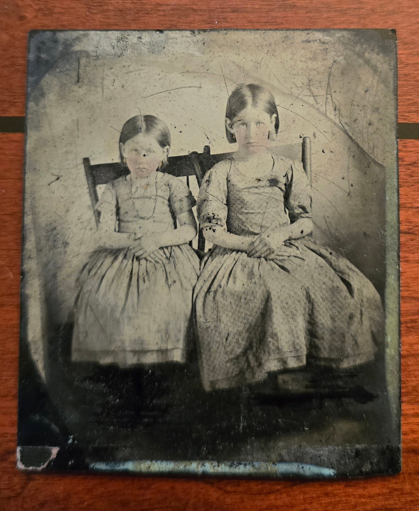 1/6th plate Ambrotype of two girls: This haunting mid-1850s–1860s 1/6th plate ambrotype captures two young sisters whose unblinking, stark gaze creates an instantly striking, eerily magnetic aura. Seated rigid and upright on simple wo