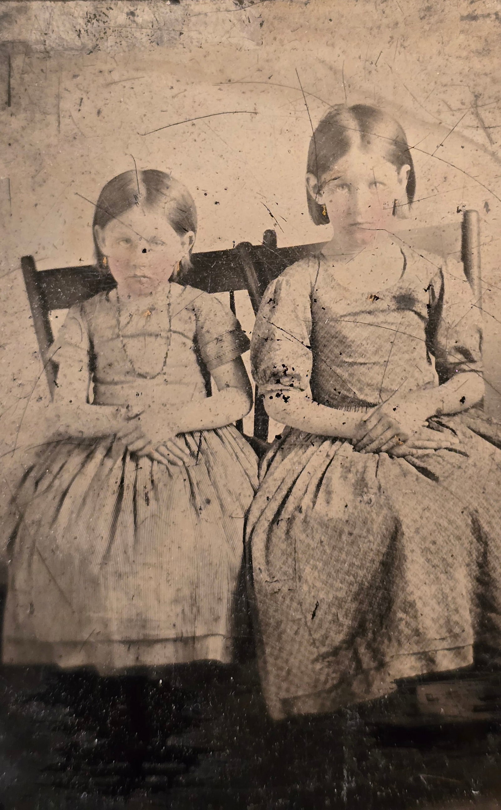 1/6th plate Ambrotype of two girls: This haunting mid-1850s–1860s 1/6th plate ambrotype captures two young sisters whose unblinking, stark gaze creates an instantly striking, eerily magnetic aura. Seated rigid and upright on simple