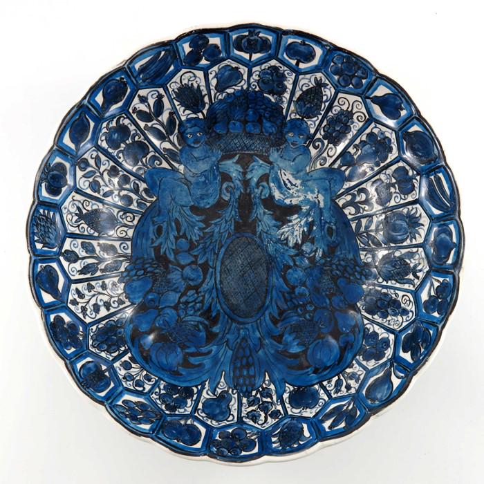 John Pearson, an Arts and Crafts painted bowl, in the: John Pearson, an Arts and Crafts painted bowl, in the Delft style, blue and white putti and fruiting festoons within a lappet border of fruit and flowers, the base with a cornucopia and monogram, 33cm