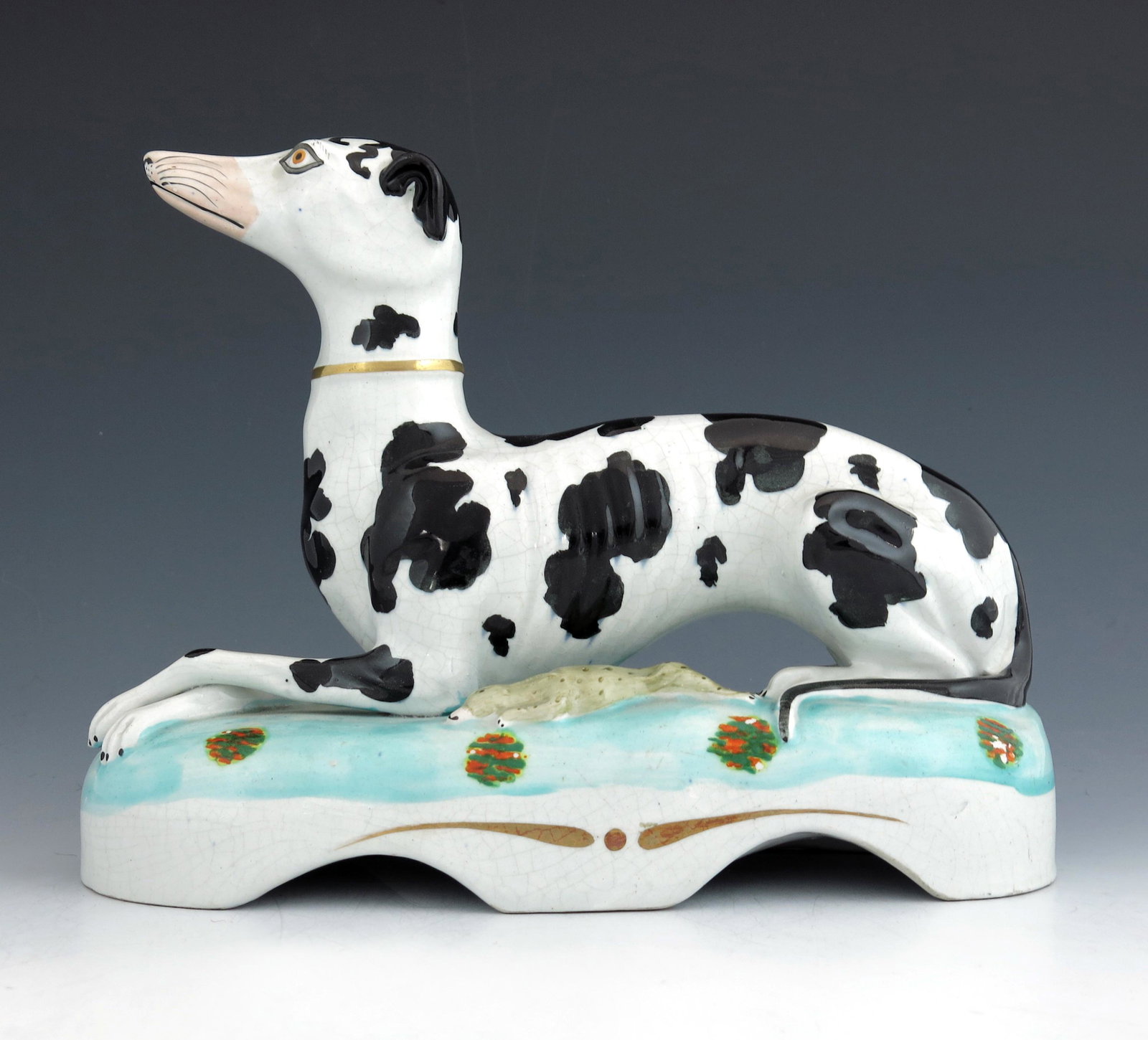 A Staffordshire pottery figure of a blac: A Staffordshire pottery figure of a black and white 'Disraeli' greyhound, circa 1860, modelled recumbent next to a hare, on a serpentine plinth base with floral painted motifs.
