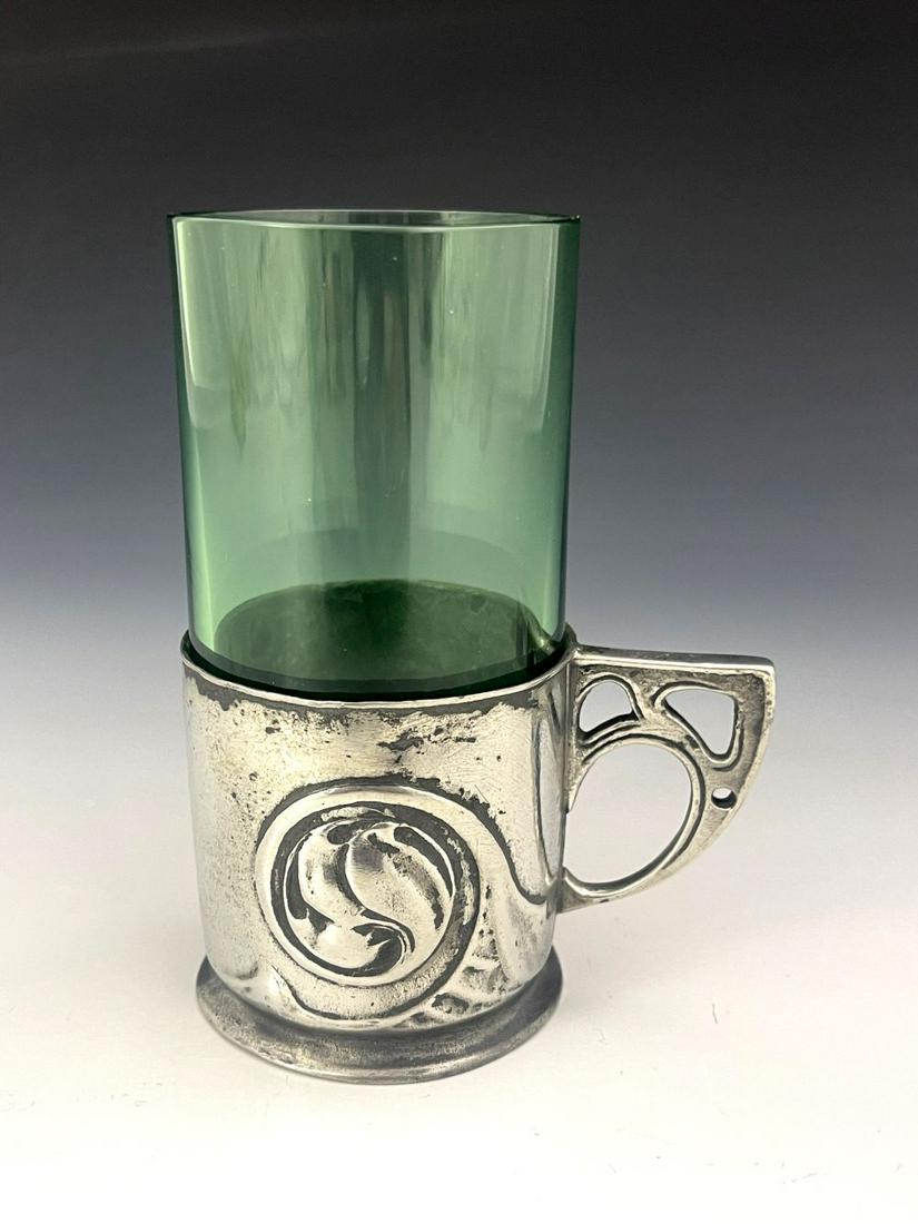 Archibald Knox for Liberty and Co., an A: Archibald Knox for Liberty and Co., an Arts and Crafts Tudric pewter and glass mug, model 0358, circa 1905, the cylindrical holder with moulded coiled bud motif, angled and pierced handle, with green