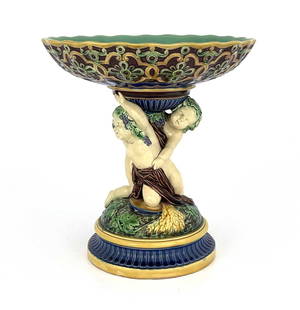 A Minton majolica figural comport, 1863,: A Minton majolica figural comport, 1863, modelled as two Bacchanalian putti on a vine moulded domed and fluted base, supporting a Renaissance trellis scalloped dish, impressed marks, 29.5cm high