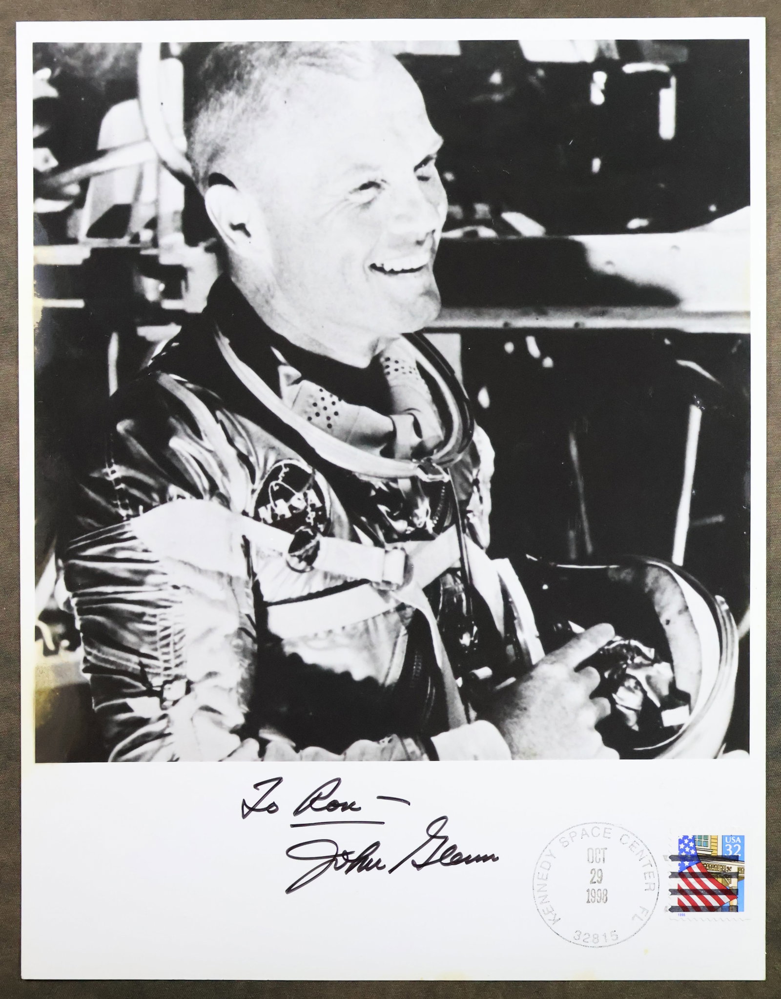 John Glenn Signed & Postmarked Photograph: This 8" x 10" black and white photograph shows a jolly Astronaut John Glenn in his space suit. The photograph has been boldly hand signed and dedicated by John Glenn. The photograph also displays the 