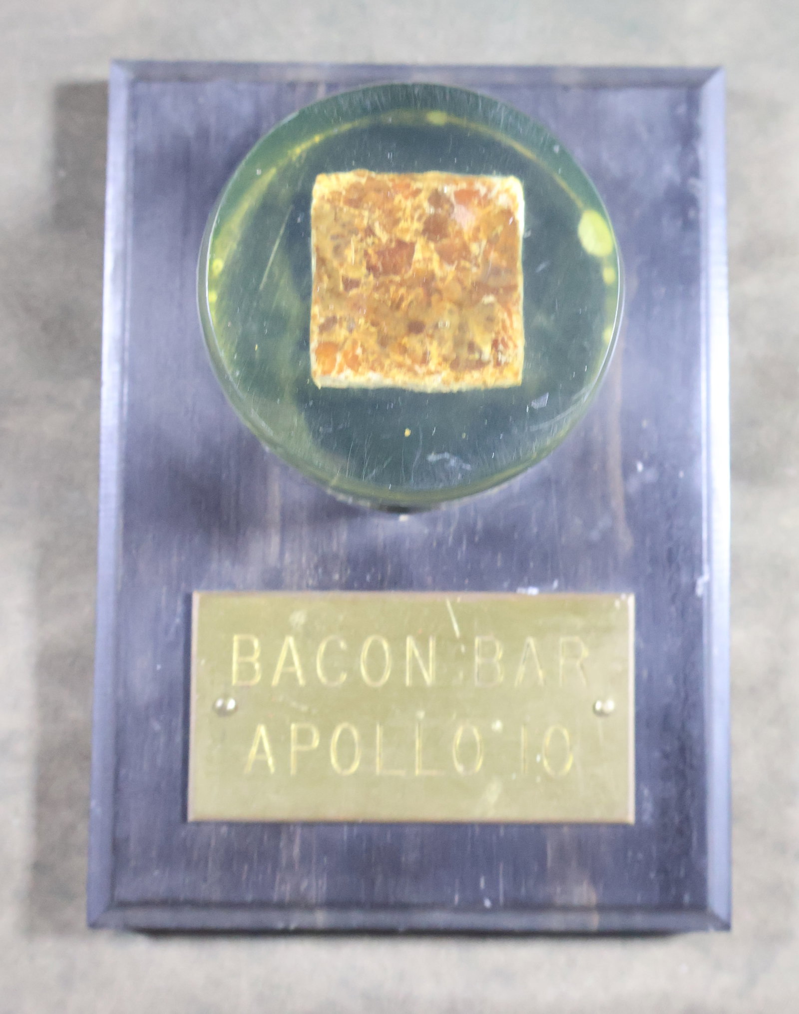 Apollo 10 Bacon Bar Lucite Display: This 5" x 4" display plaque/desk showcases a 2" x 1" round Lucite containing a 1" square section of Bacon Bar, a space food item that was part of the menu items aboard the Apollo 10 mission. The