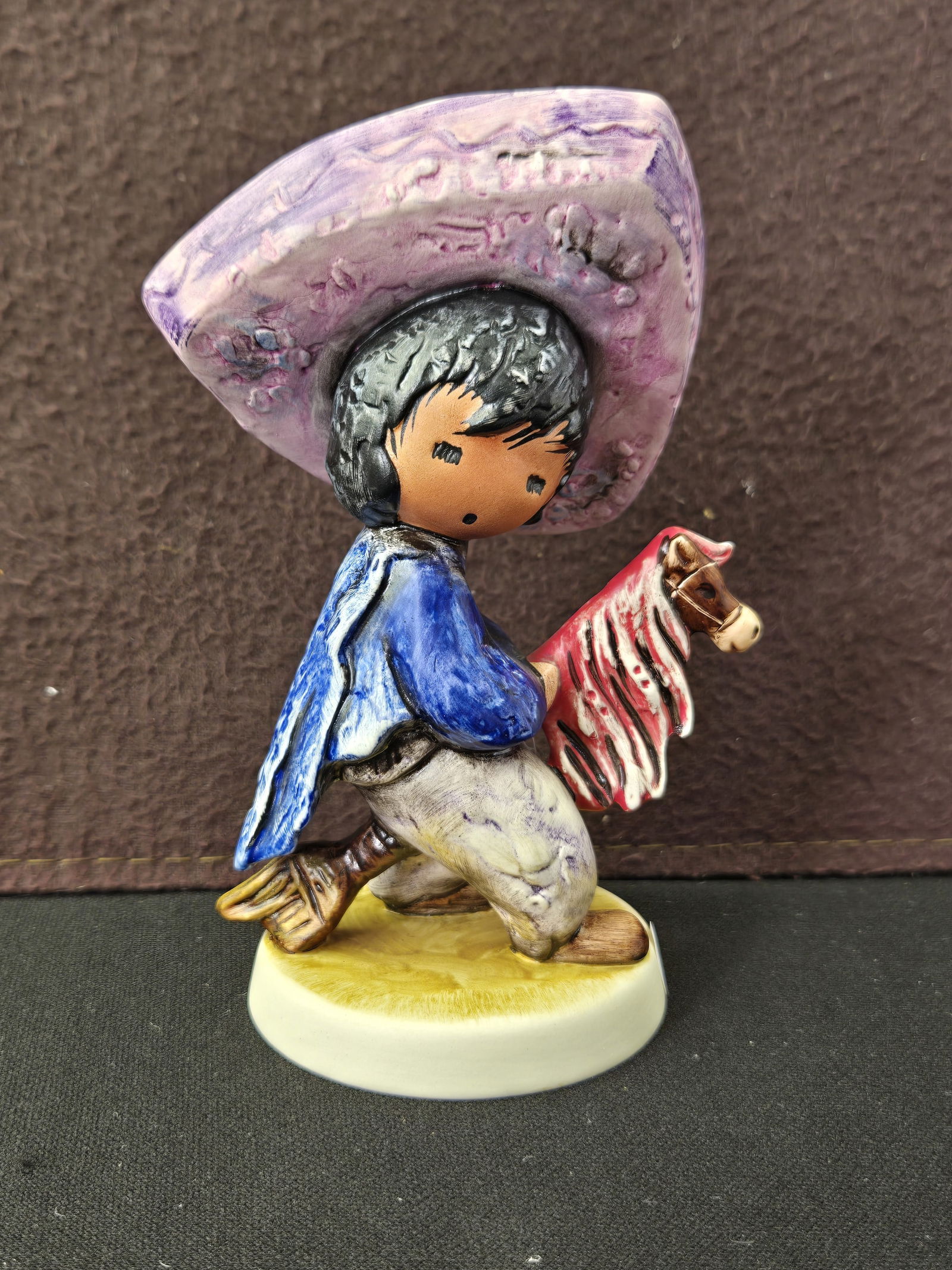 1983 Goebel DeGrazia 'My First Horse' IO 312 Figurine: This listing features a 1983 Goebel DeGrazia 'My First Horse' IO 312 ceramic figurine. The hand-painted piece depicts a child in a prominent purple sombrero and blue top, kneeling while "riding" a red
