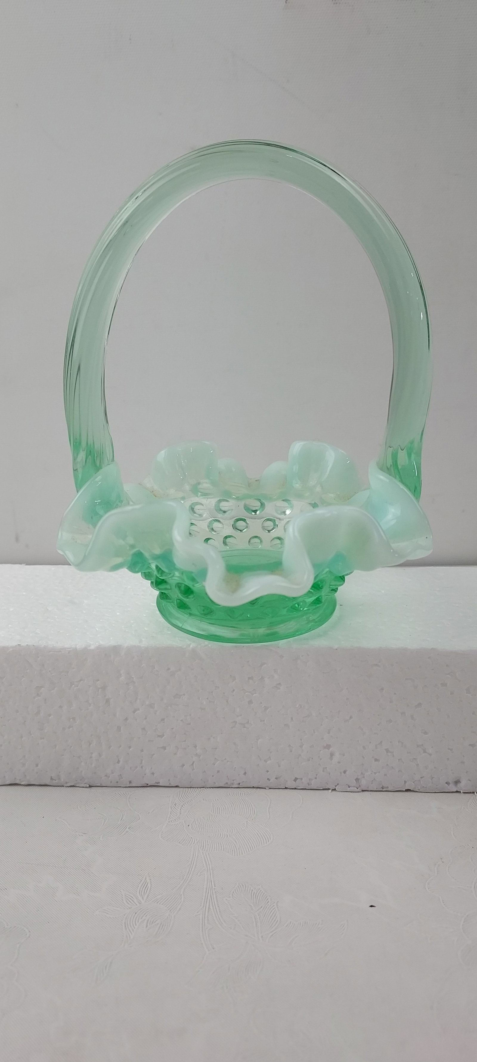 Fenton Art Glass Willow Green Opalescent Hobnail Basket: A vintage Fenton Art Glass basket crafted from Willow Green Opalescent glass. The piece features a distinct hobnail pattern on its base, complemented by a ribbed, C-shaped handle applied to the rim. T