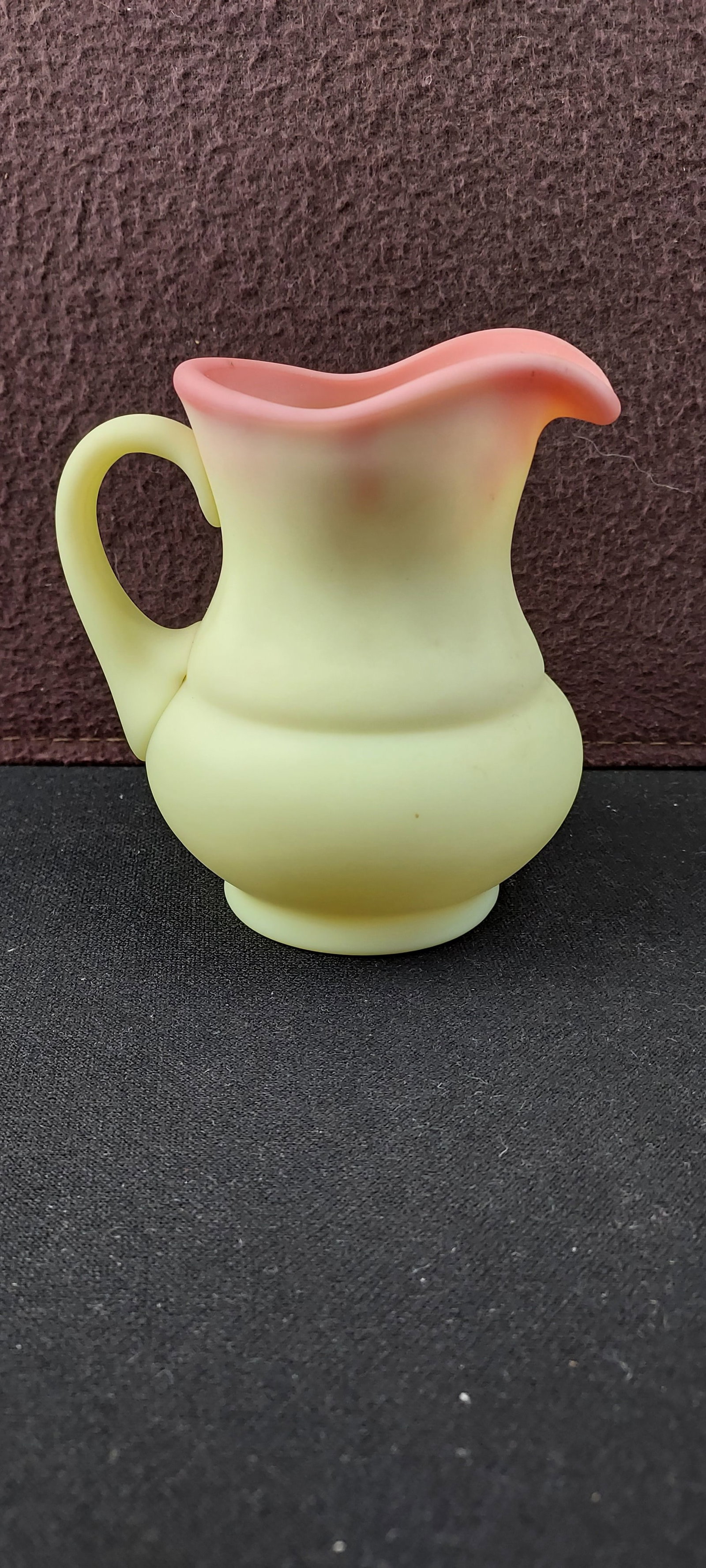 Fenton Burmese Glass Creamer: This listing features a classic Fenton Burmese glass creamer. The item is a small, loose glass pitcher or creamer, distinguished by its iconic two-tone color gradient. The body exhibits a pale yellow-