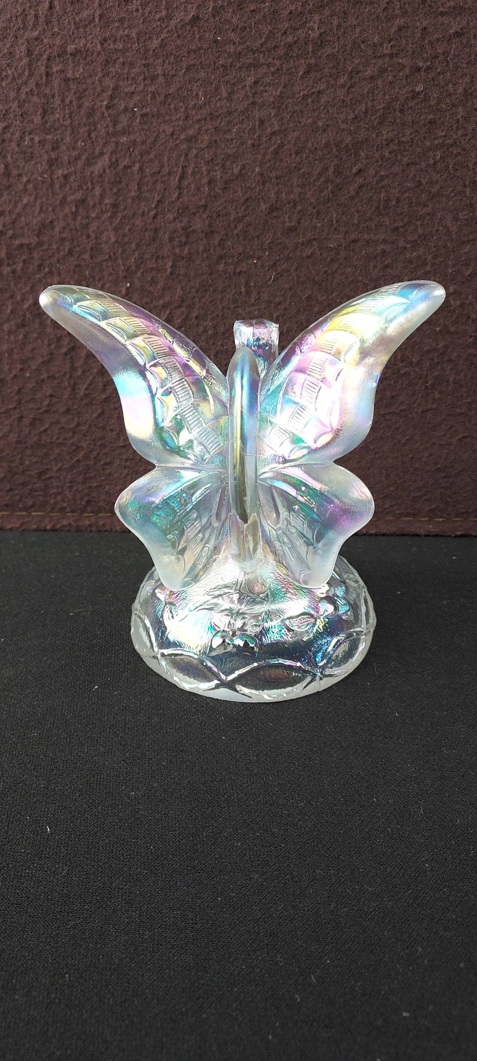 Fenton Iridescent Butterfly on Branch: A Fenton Art Glass figurine depicting a butterfly with outstretched, patterned wings, resting on a circular, relief-molded base adorned with a leaf or floral motif. The piece is crafted from clear gla