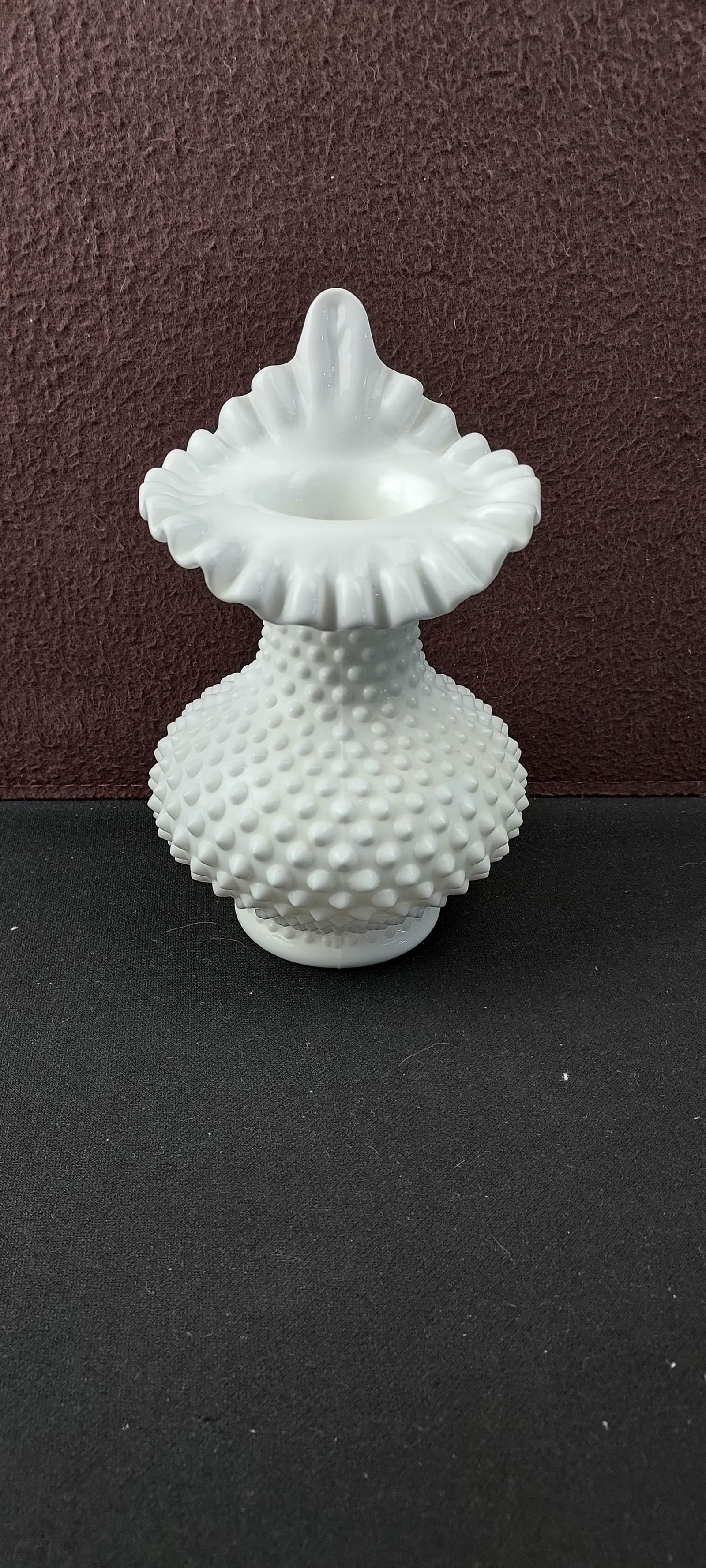 Vintage Fenton White Milk Glass Hobnail Ruffled Crimped Vase: A vintage Fenton white opaque milk glass vase, featuring a bulbous body adorned with a textured hobnail pattern. The body tapers to a narrower neck, which culminates in a prominently ruffled and crimp