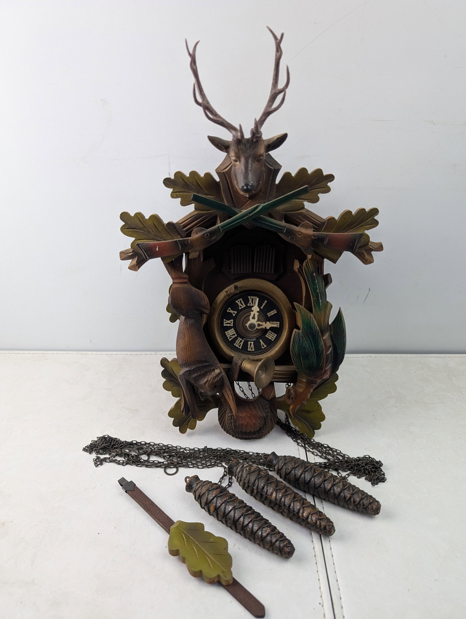 Vintage West German Schmeckenbecher Hunting Theme Cuckoo Clock: A vintage West German cuckoo clock, featuring a traditional carved wooden case with a prominent hunting theme. The design includes a deer head with antlers at the apex, flanked by crossed rifles and o
