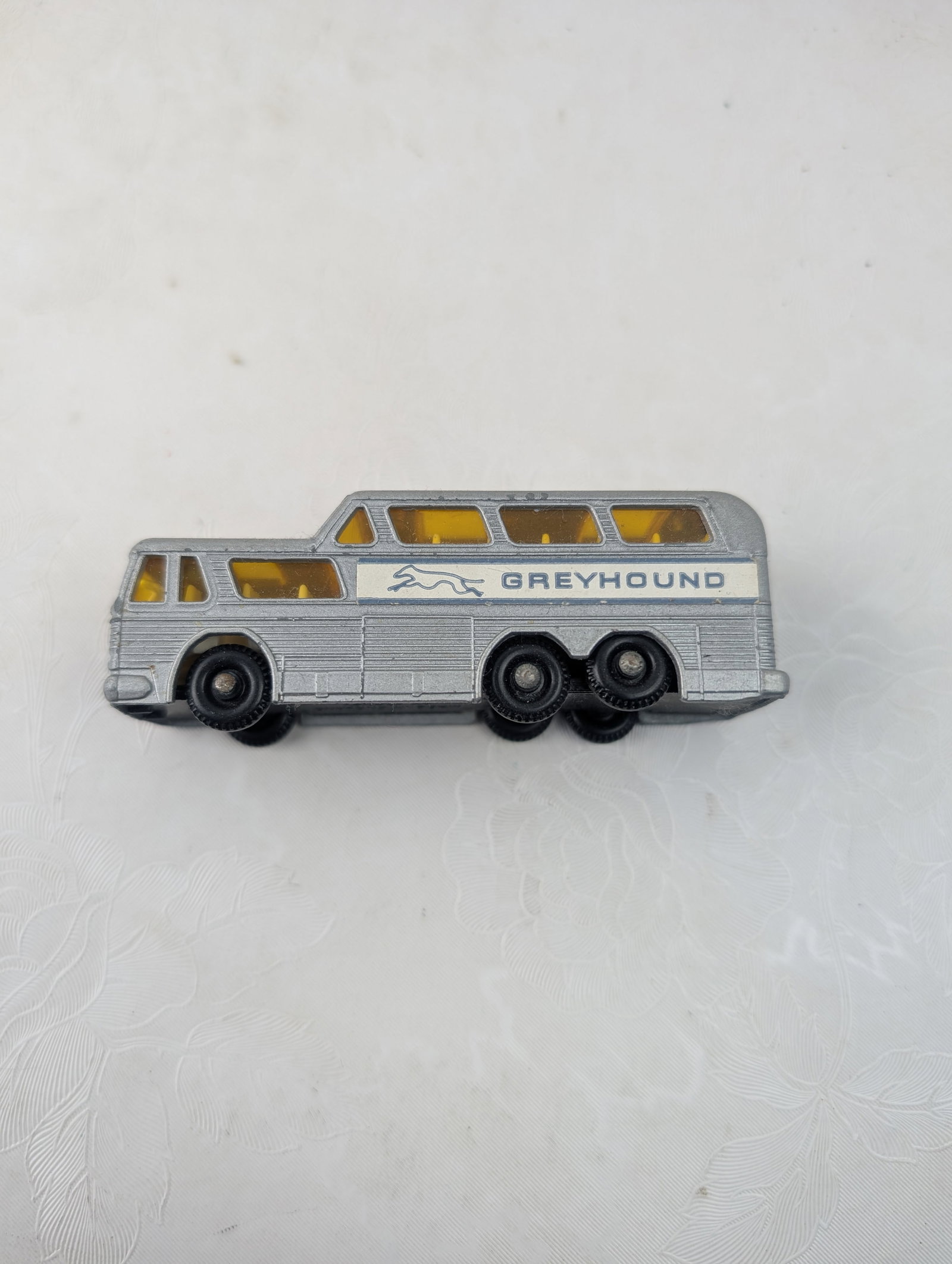 Vintage 1967 Lesney Matchbox No. 66 Greyhound Coach: A vintage Lesney Matchbox Series No. 66 Greyhound Coach, likely produced circa 1967. This die-cast toy vehicle features a silver-painted body with transparent yellow window inserts. The black base is