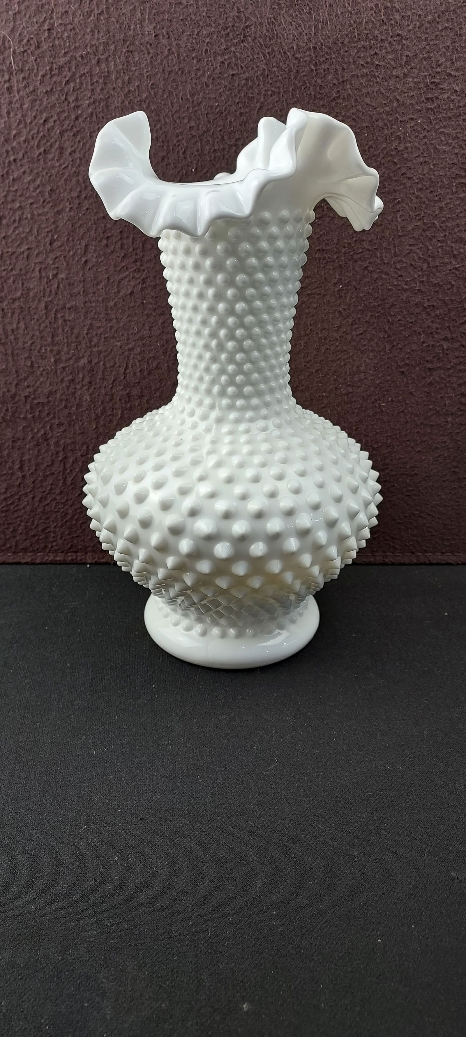Fenton White Milk Glass Hobnail Ruffled Vase, Approximately 11 Inches: This lot features a vintage Fenton White Milk Glass Hobnail Ruffled Vase. The vase is crafted from opaque white milk glass, characterized by a distinct hobnail pattern covering its bulbous body and ta