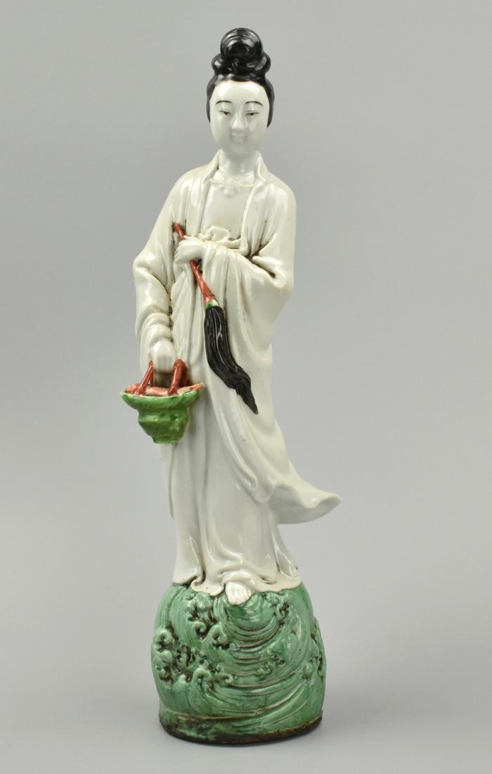 Chinese Painted Figure of a Woman,ROC Period: A Chinese, Dehua porcelain figure of a dark-haired woman in a windswept robe standing on a circular plinth of green waves, with a copper-red & green basket of fish in one hand, a long bristled copper-