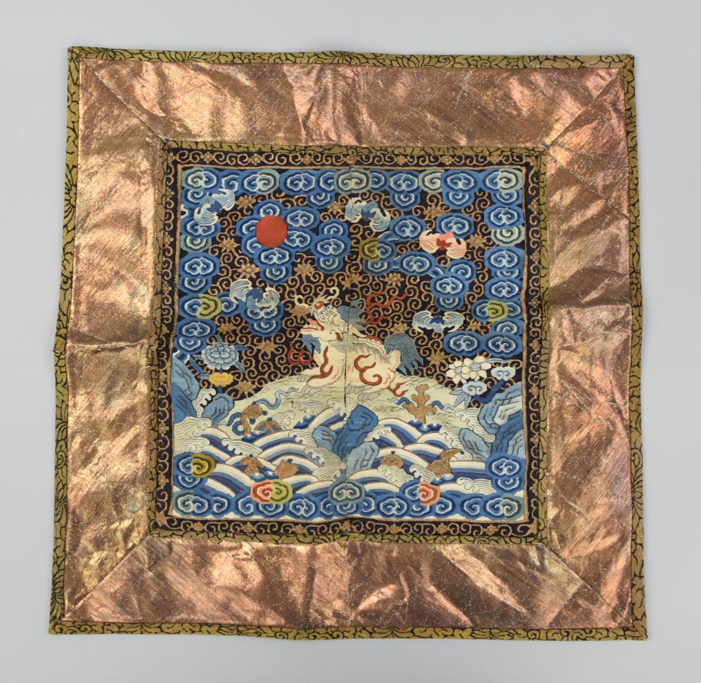 Chinese Military 2nd Rank Lion Buzi, Qing Dynasty: A Qing Dynasty badge of second degree military rank with a Buddhist lion seated above a series of waves with clouds & bats against a black & gold scrolling sky above, this bordered by further black & 