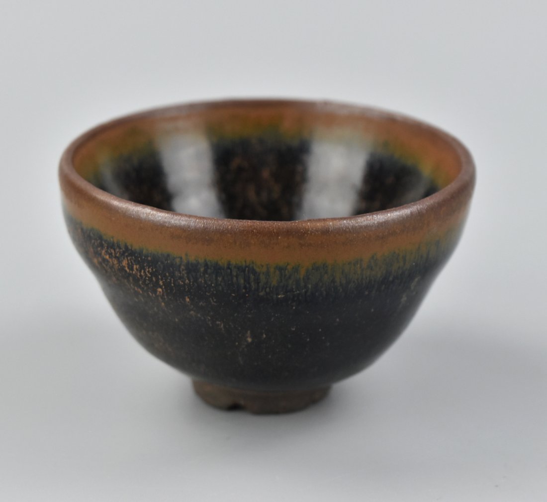 Chinese Jian Ware Hare`s Fur Tea Bowl, Song: the steep sides rising to a shallow groove below the gently everted rim, covered overall with a black glaze suffused with fine russet striations,the warm orange amber tone concentrating around the rim