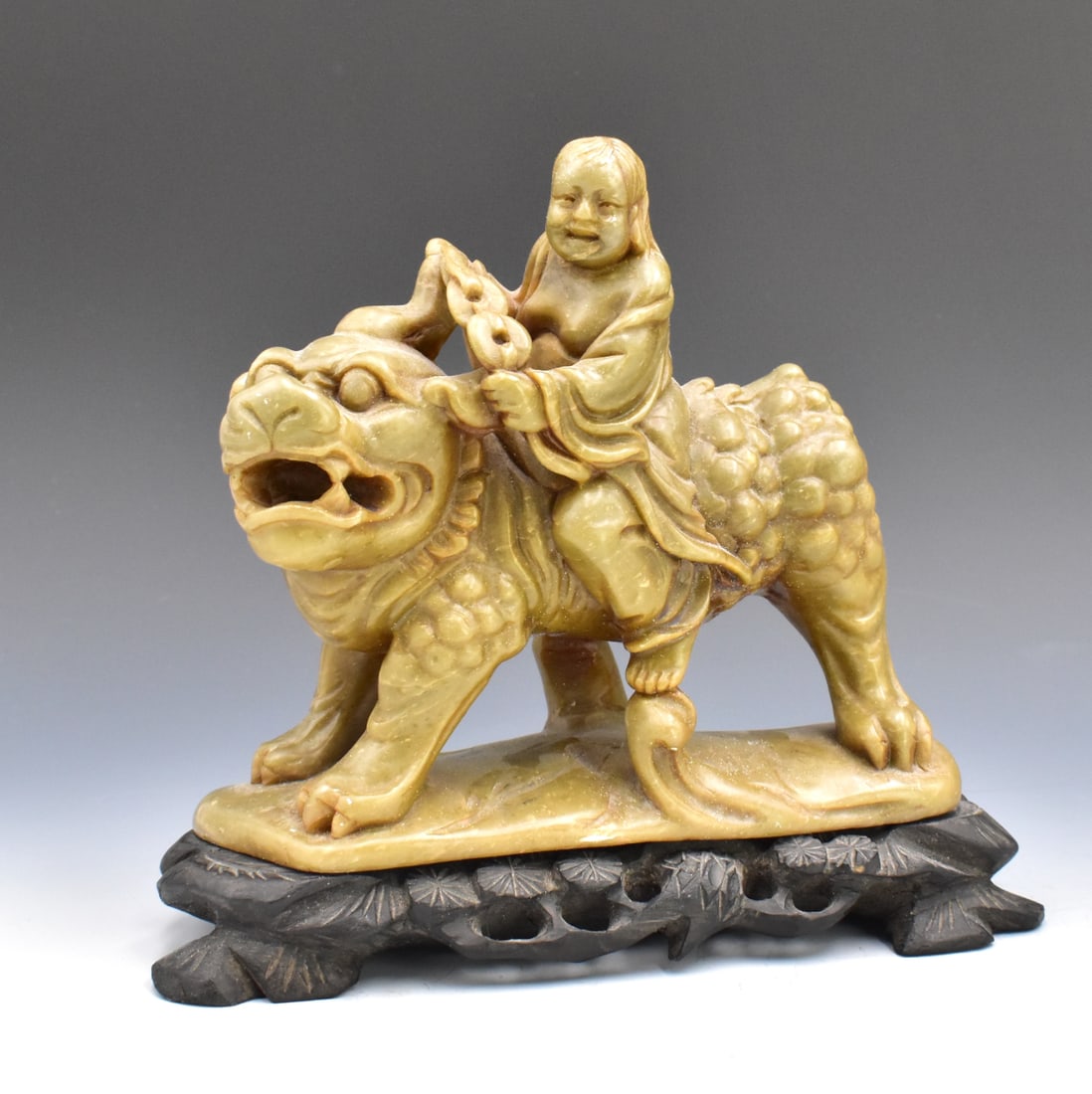 Chinese Soapstone Carved Figure on Kirin: A Chinese soapstone carved boy riding a Kirin beast, over a carved wooden base. Boy holds the beautifully carved reigns and turns the beast to his left. He is smiling and in control. D. 6" (15.2 cm) L