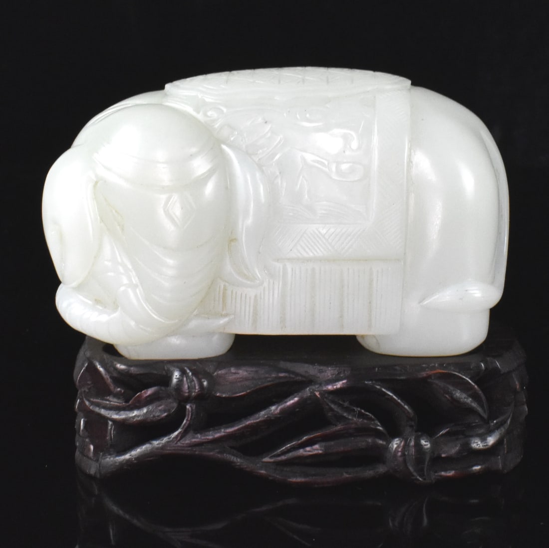 Chinese White Jade Carved Elephant on Stand: A Chinese white jade carved elephant on wooden stand. A thick rounded form with incised head facing to its left, an ornate blanket saddle draped over the body. With a fitted wooden stand with deeply i