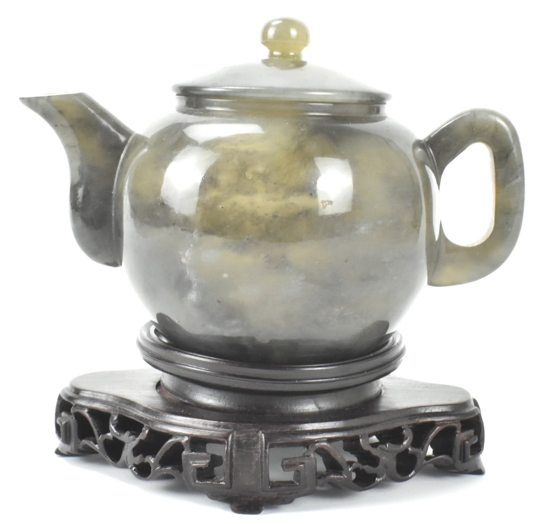 Chinese Hard Stone Carved Teapot on Wooden Stand: A Chinese hard stone carved teapot on a wooden stand. A smooth round teapot with C shaped handle and curved spout. The cover extends beyond the neck, and spout is wide, short, and curved. Dark grey st