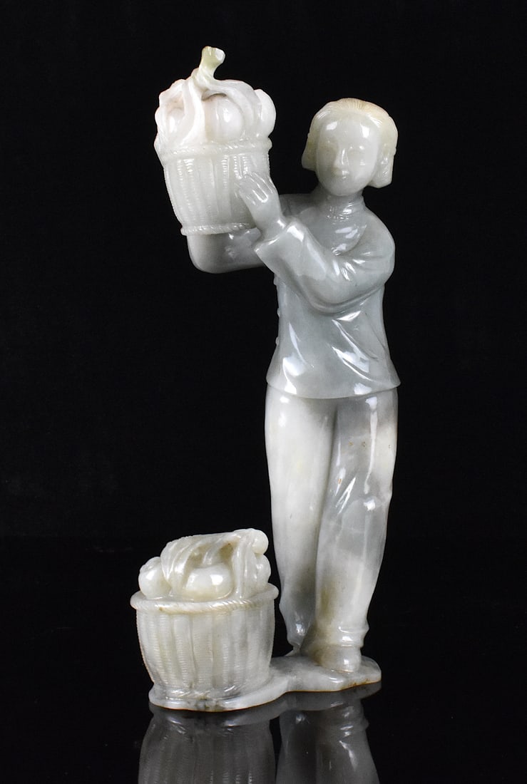Chinese Jadeite Carved Lady Holding Fruit Basket: A Chinese jadeite carved lady holding fruit basket. The lady stands holding a basket of fruit aloft with both hands to her right. Another basket in carved jadeite is on the ground below her. H. 8 1/4"