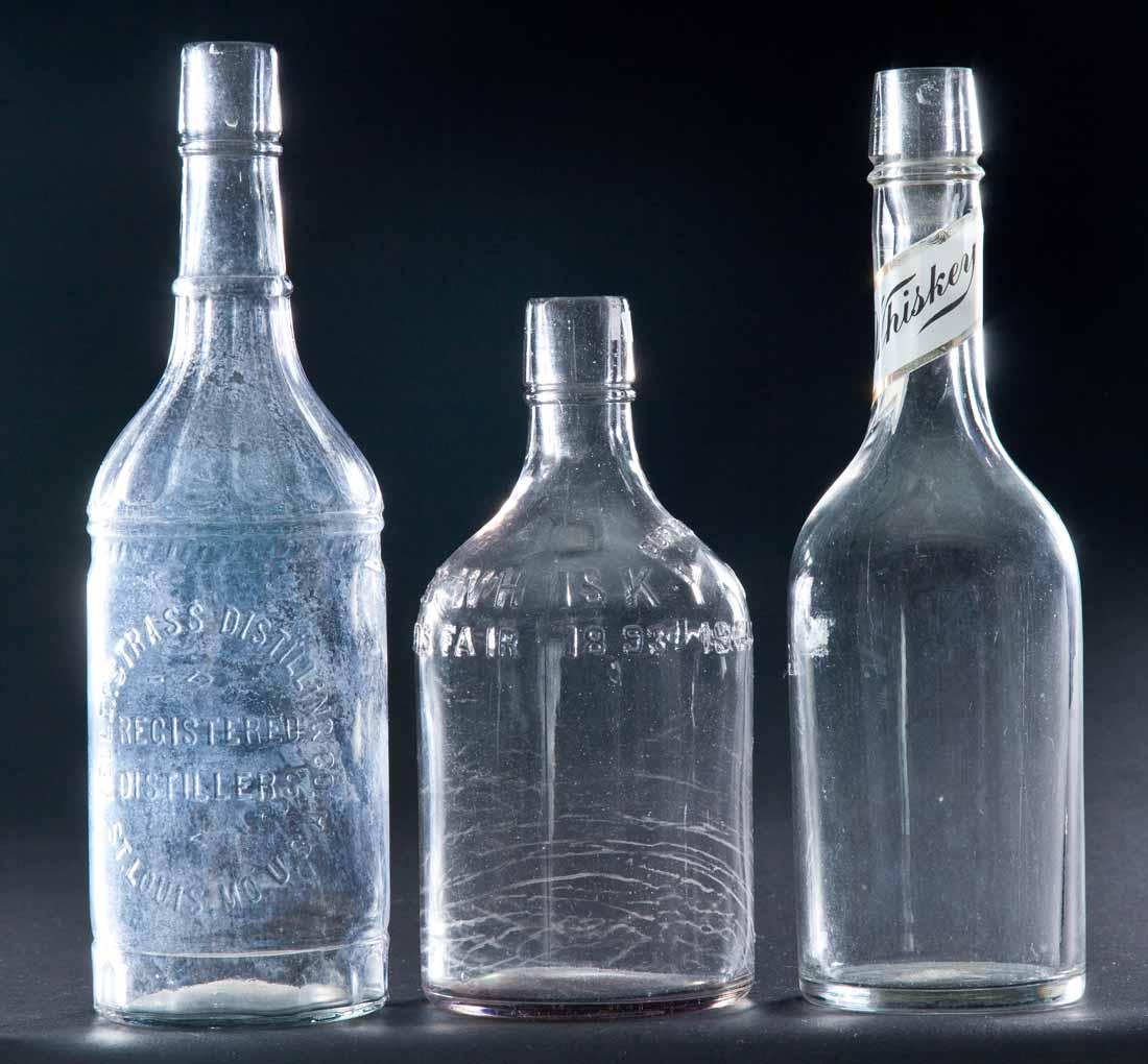 THREE COLORLESS WHISKEY BOTTLES: All pre-Prohibition. Blown with label under glass "Whiskey". 11.25"h. One with embossed label, "Old Time's Whiskey, Grand Prize World's Fair, 1893 1904". 8.75"h. One with embossed label, "Kellerstrass